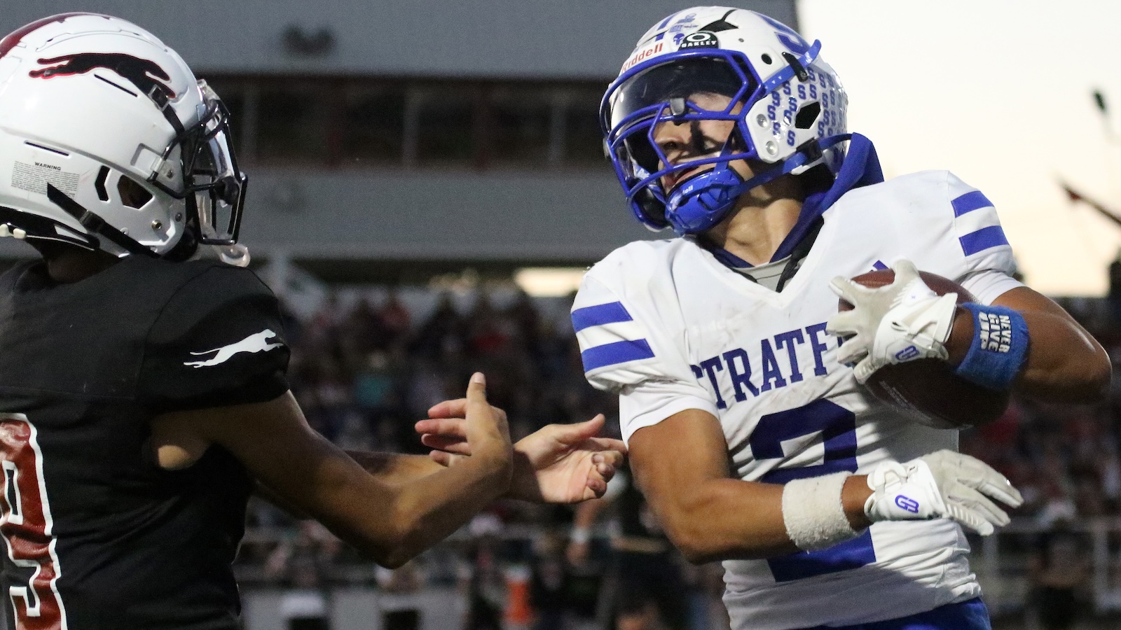 Football players in a game: a white-and-blue helmeted player clutches a football while fending off a tackler in black uniform.