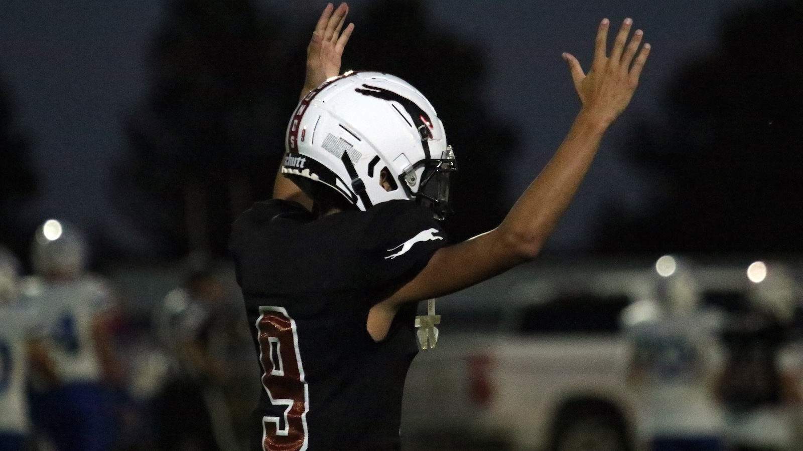 Football player with arms raised in celebration, wearing a white helmet and black jersey number 3 on the back, on a dusk field.