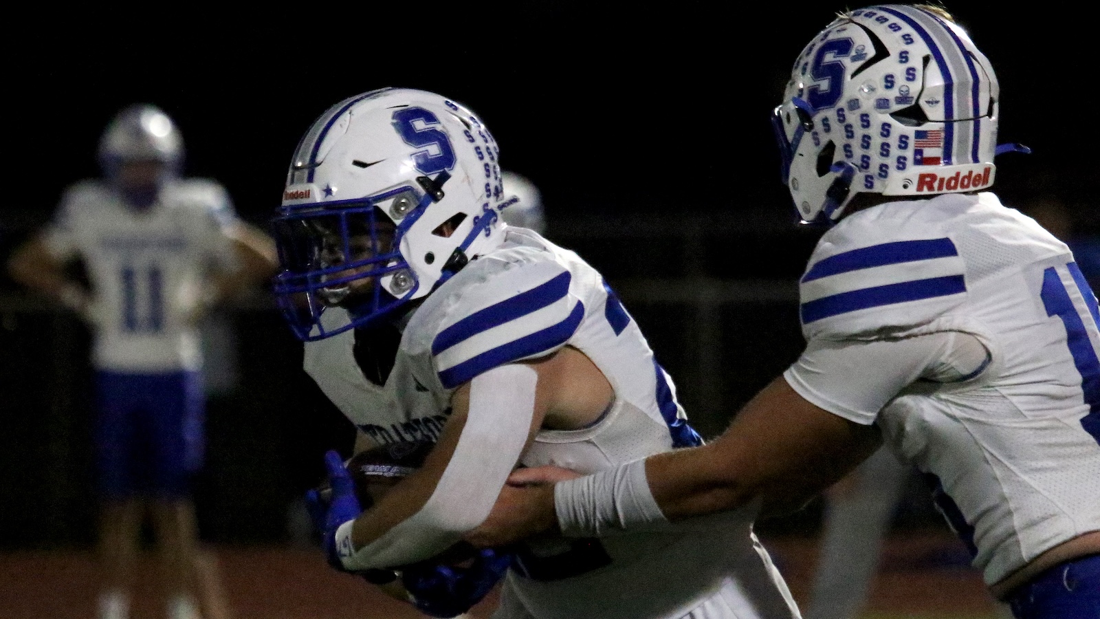 Two football players in white jerseys with blue stripes tackle each other during a night game on the field.