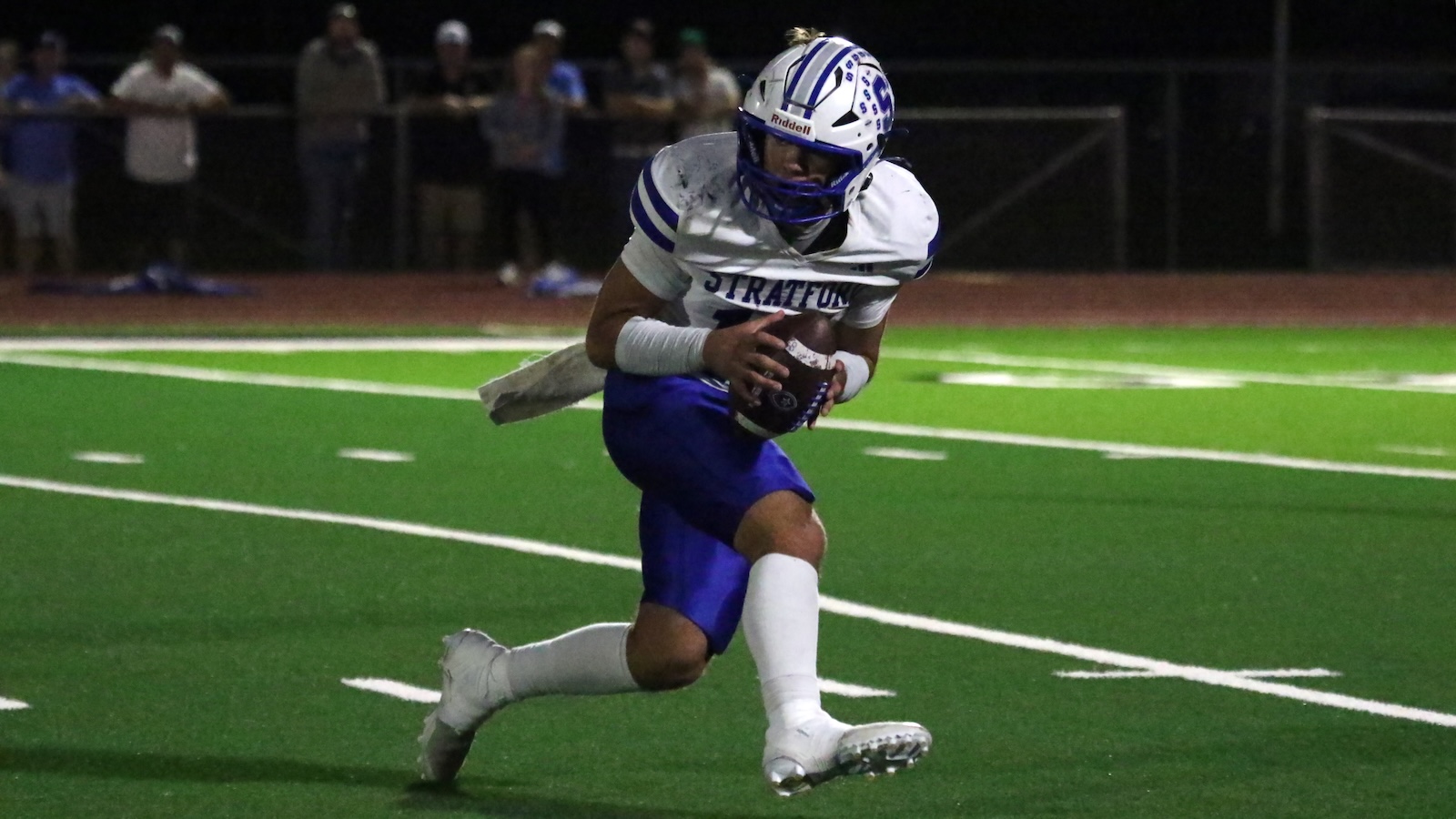 Night game action: a Stratford football player in white and blue runs with the ball on a bright green turf field.