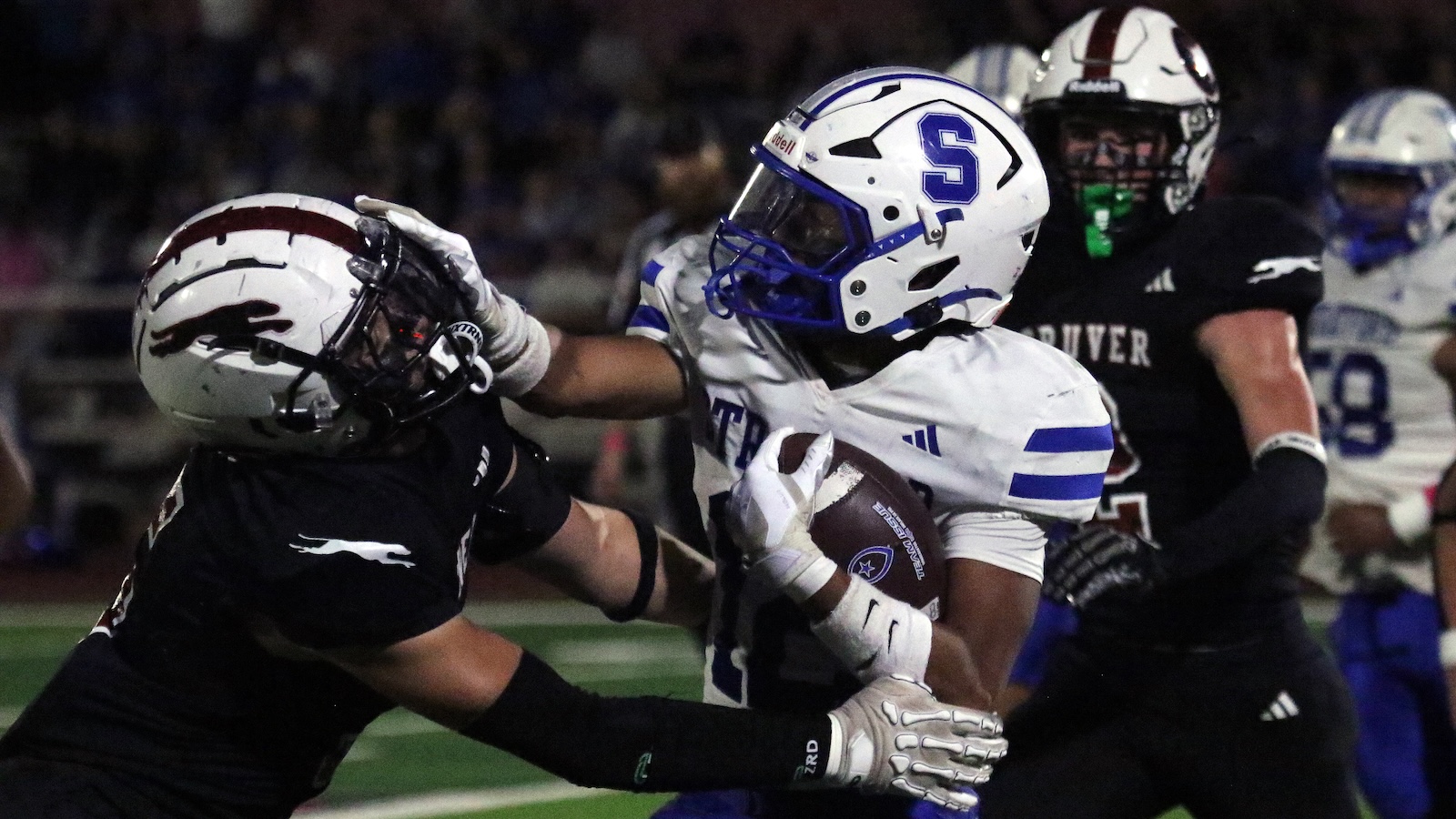 White-jersey football player runs with the ball as a defender in black reaches in for a tackle and stops him short of a gain.