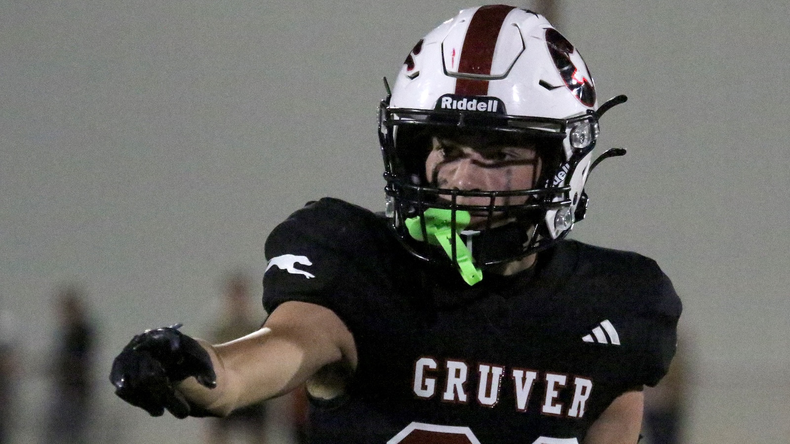 Gruver football player in a black jersey reaching forward, white helmet with red stripe and neon green mouthguard during a game on an indoor field.