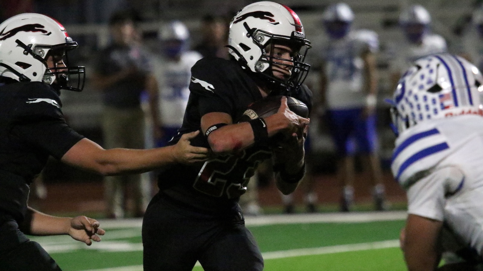 Football player in black runs with the ball as a tackler grabs his jersey during a night game on a green turf field with defenders nearby