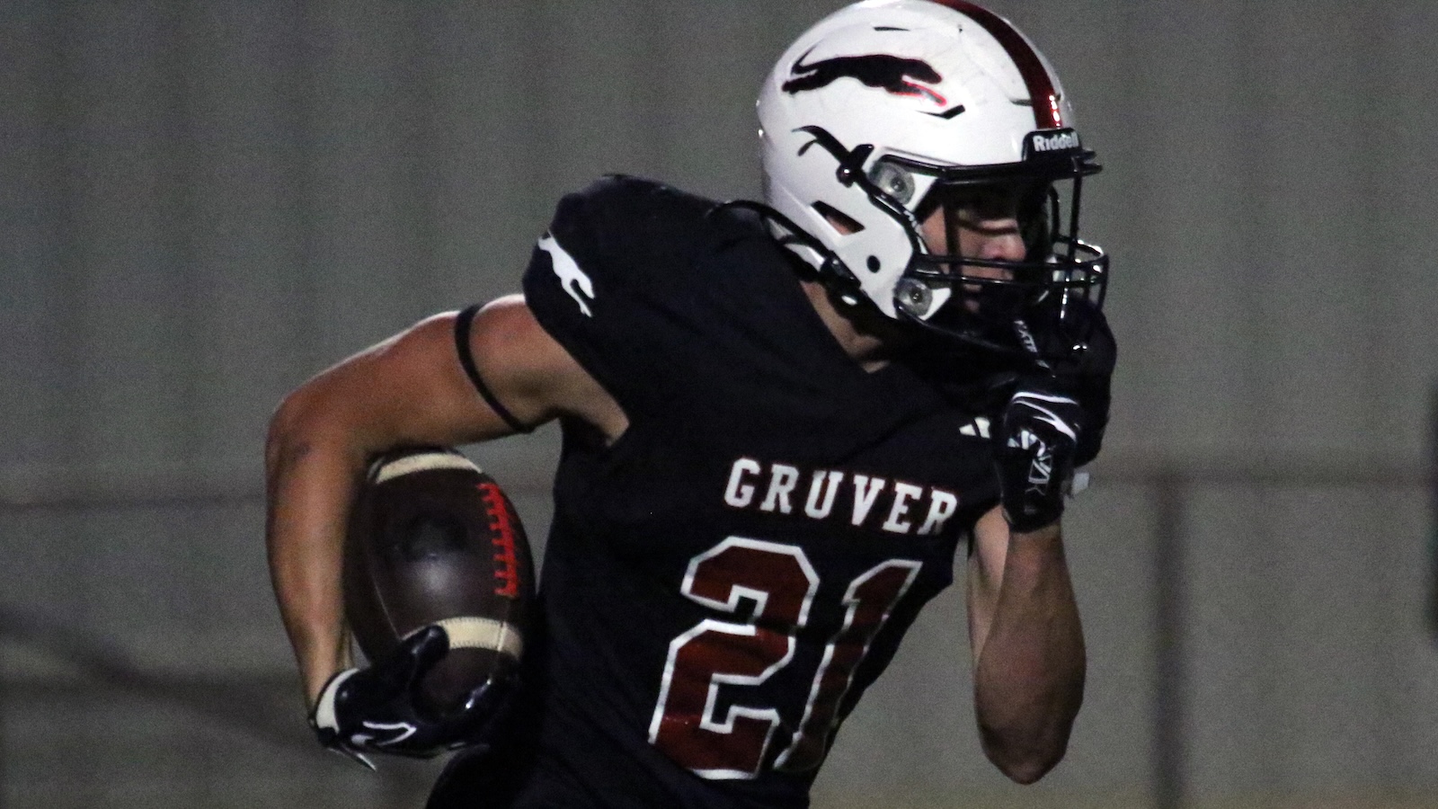 Football player in a black Gruver jersey with number 21 runs with the ball during a night game, wearing a white helmet and gloves and looking ahead.