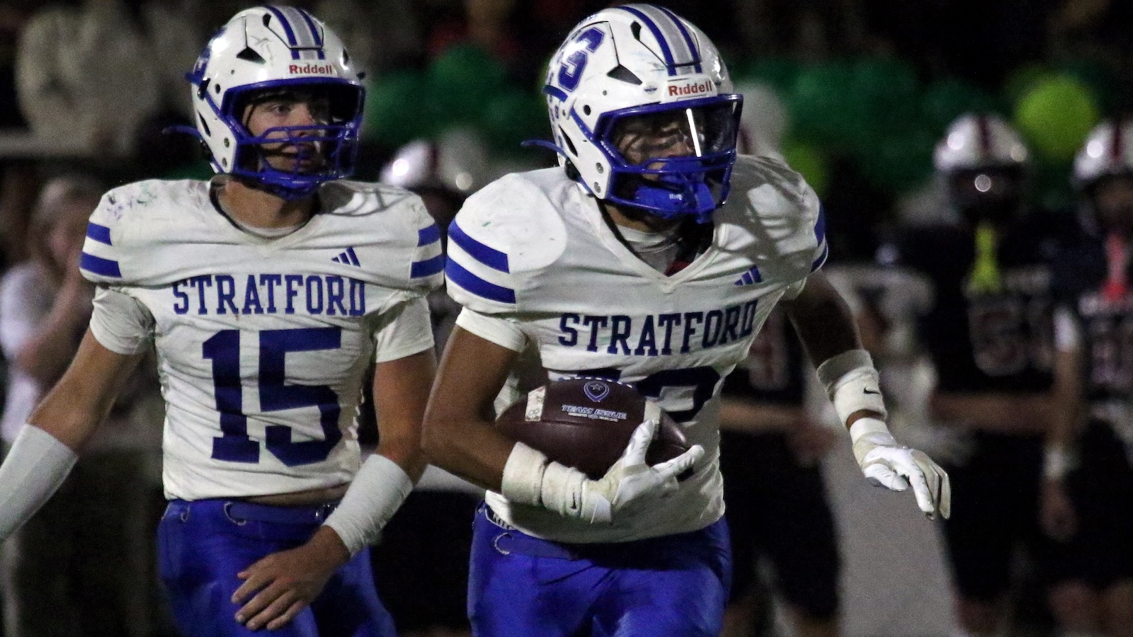 Two Stratford football players in white and blue uniforms run at night, with one carrying the football as his teammate runs beside him.
