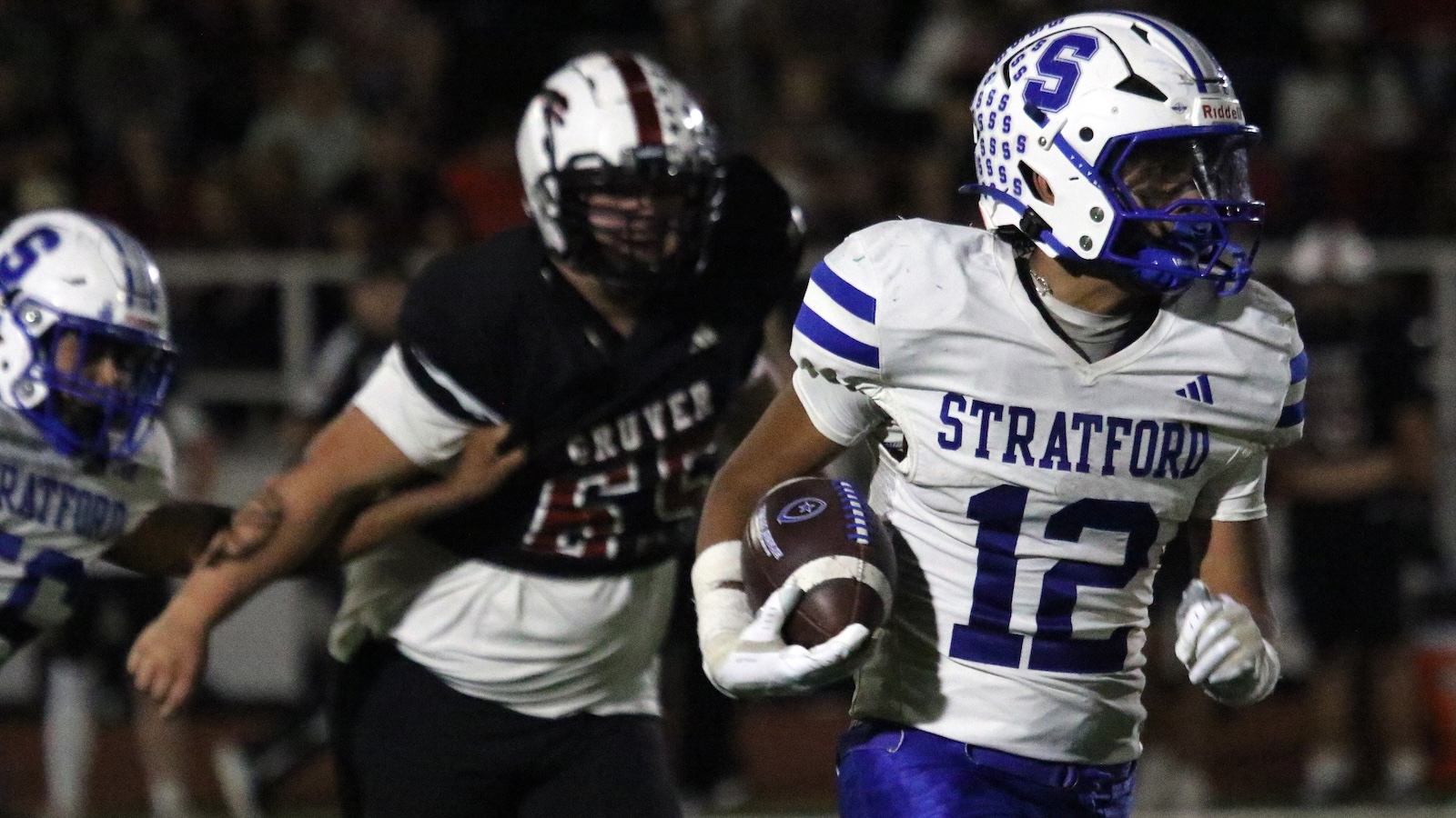Stratford player wearing white jersey number 12 runs with the football during a night game, pursued by a defender in black.