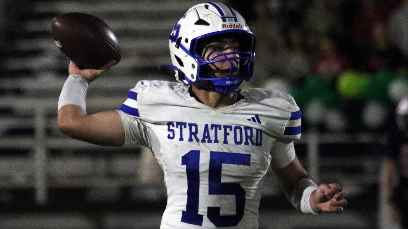 Stratford quarterback #15 throws a football during a night game in a white-and-blue uniform