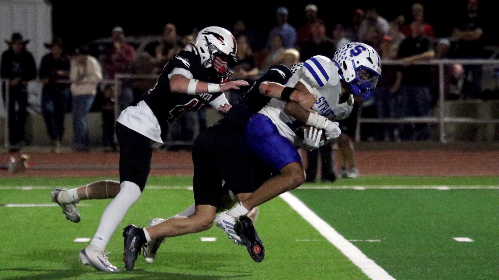 Two football players collide during a nighttime tackle on a green turf, with spectators in the background.
