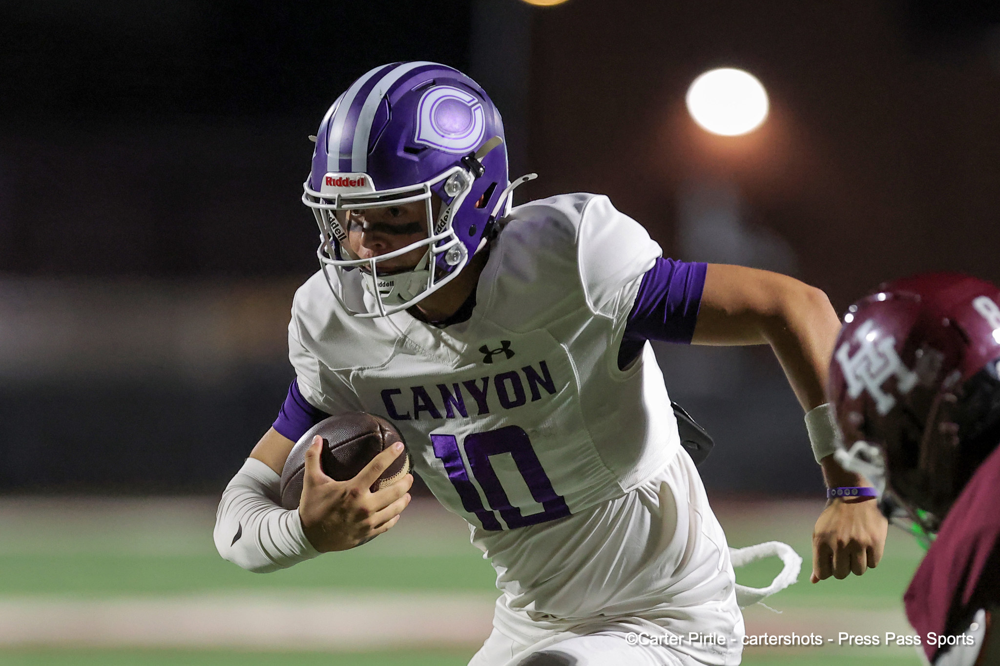 Canyon football player wearing a purple helmet and white jersey runs with the ball during a night game.