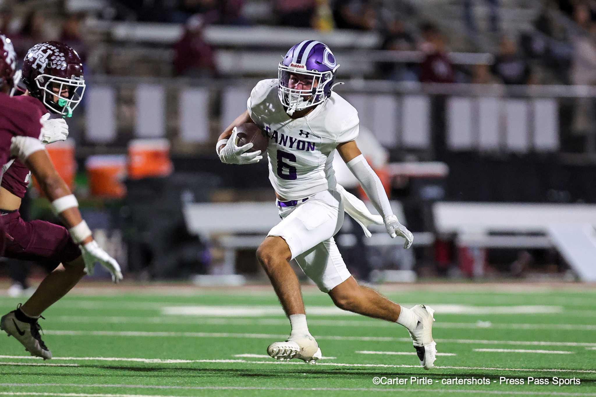 Football player in a white Canyon jersey (#6) with a purple helmet runs with the ball, chased by a maroon-uniform defender on a green turf field.