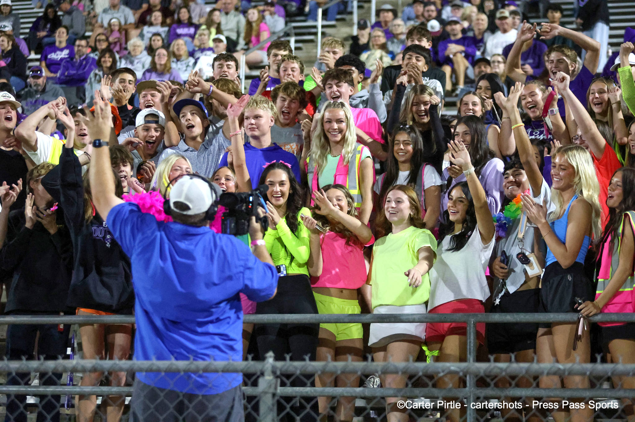 Crowd of students in bright attire cheering at a stadium, with a photographer in a blue jacket shooting from the front.