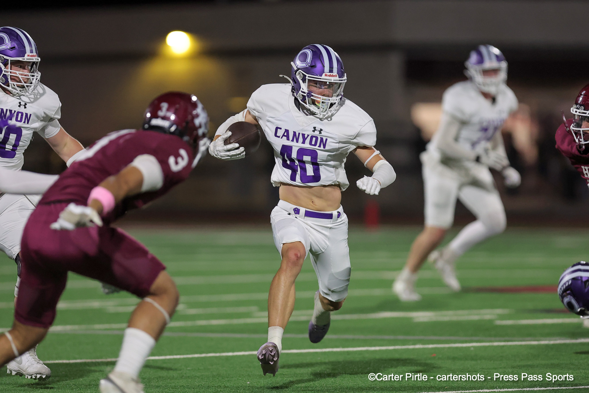 Canyon football player in white and purple runs with the ball during a night game, pursued by maroon-clad defenders.