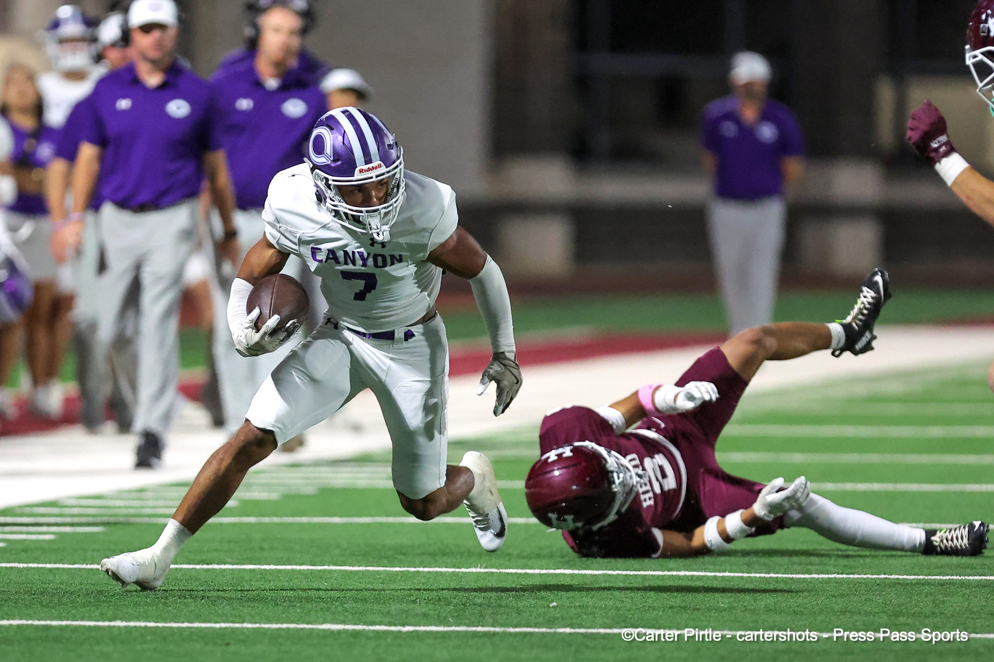 Canyon football player wearing white, number 7, runs with the ball as a maroon-clad defender dives nearby on a green turf field.