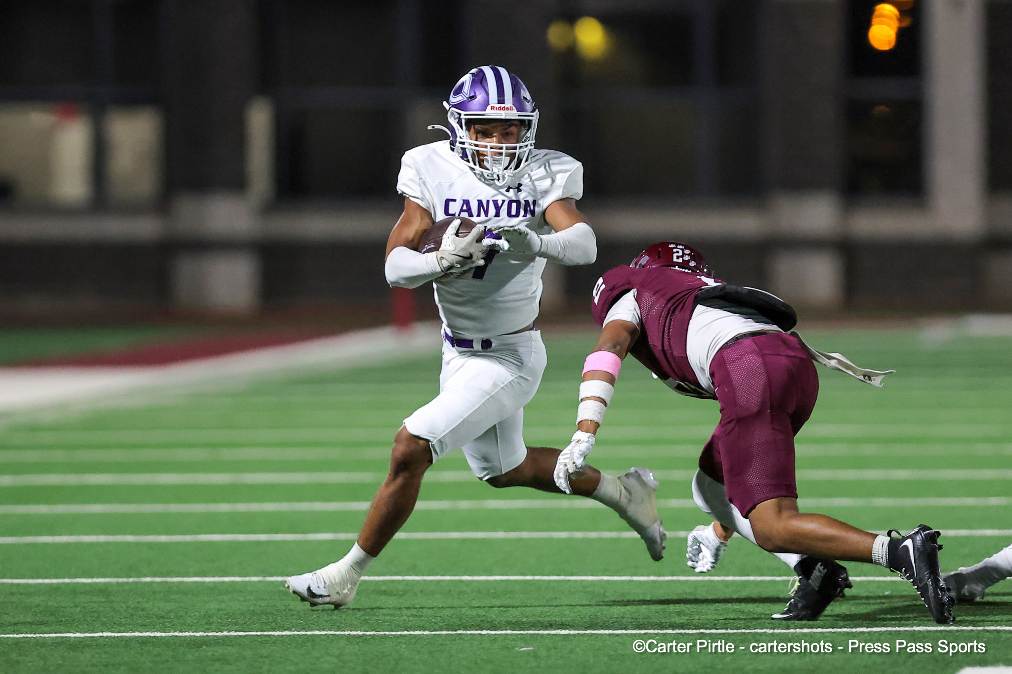 Canyon football player in a white jersey and purple helmet carries the ball as a maroon-clad defender dives in from the right on a green field at night.