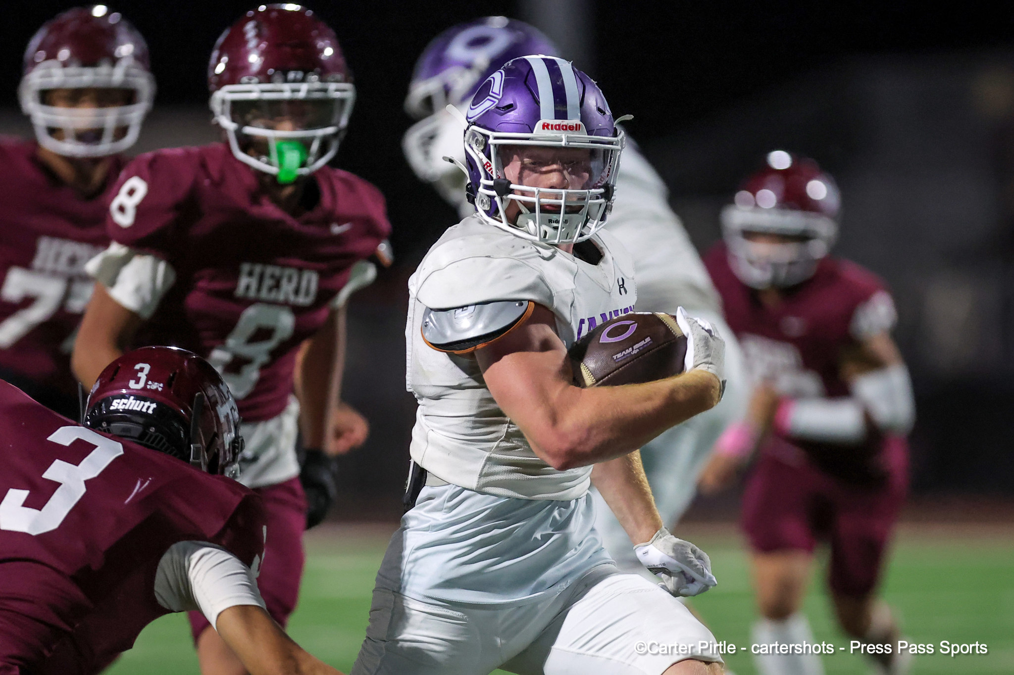 Running back in a white uniform carries the football while maroon-clad defenders pursue during a nighttime football game.