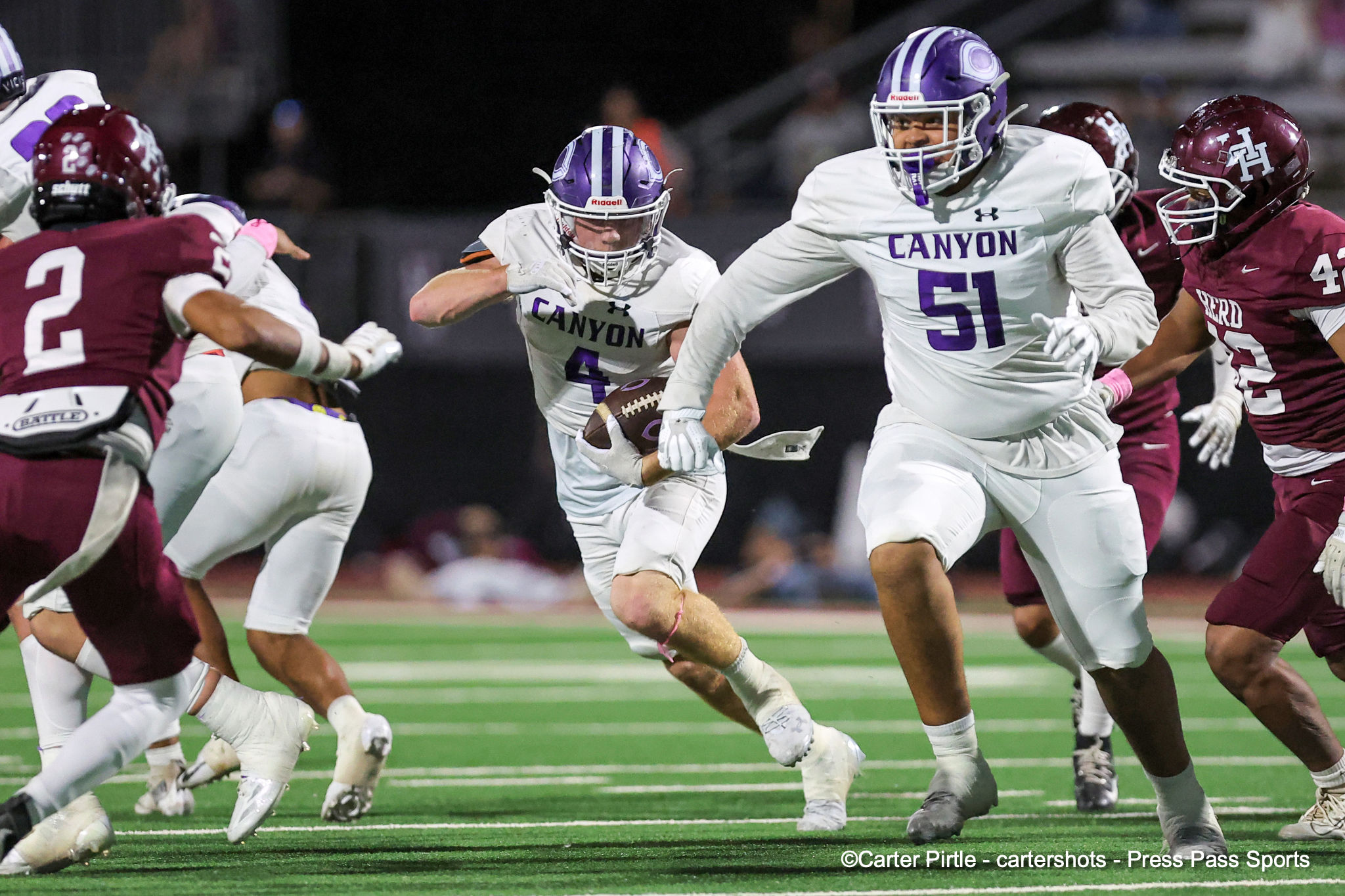 Football players in white Canyon jerseys sprint on a green turf at night, pursuing the ball carrier as maroon-clad defenders close in.