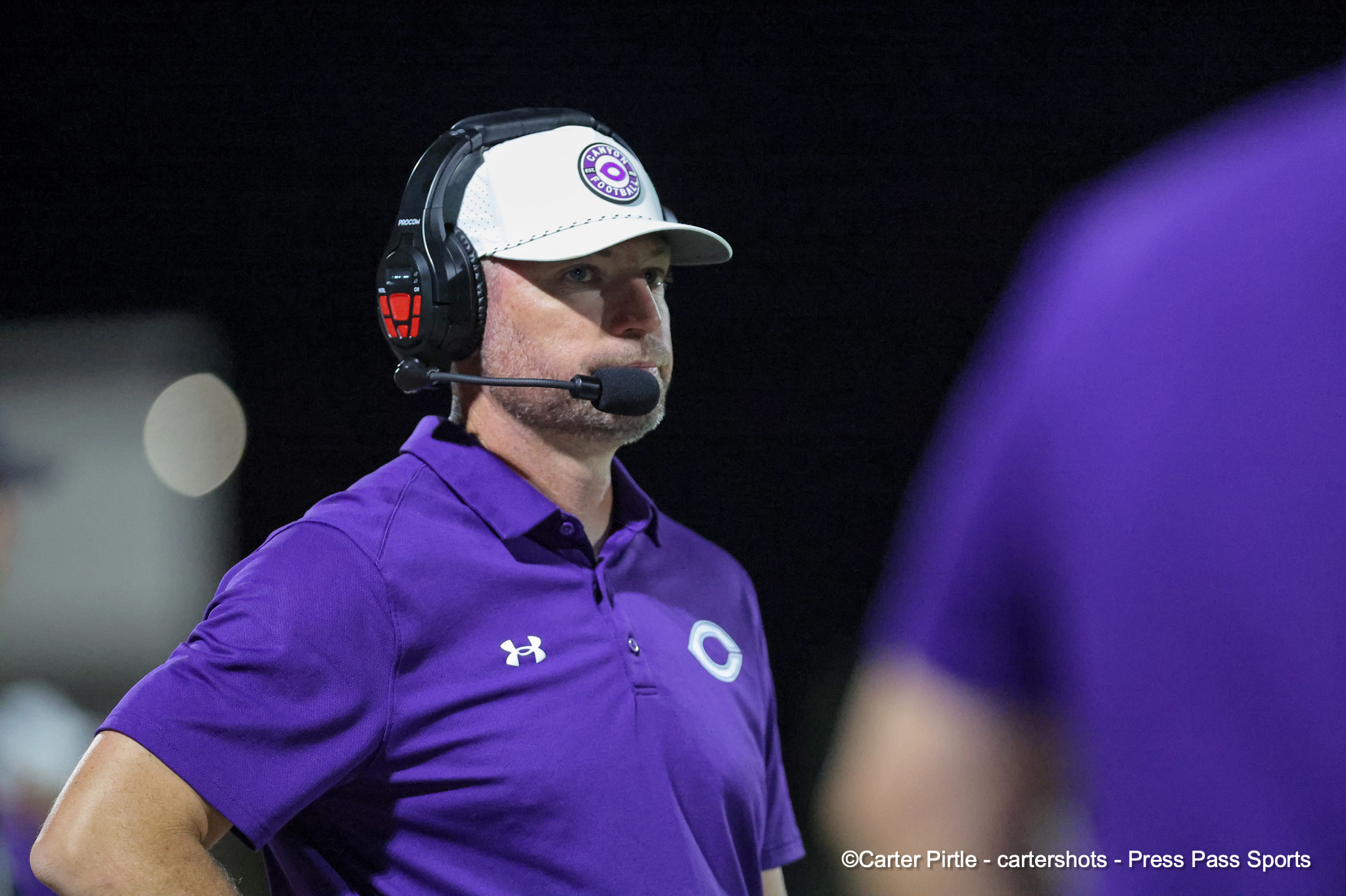 Coach wearing a headset and microphone, purple polo shirt, white cap with a circular logo, on a dark background.