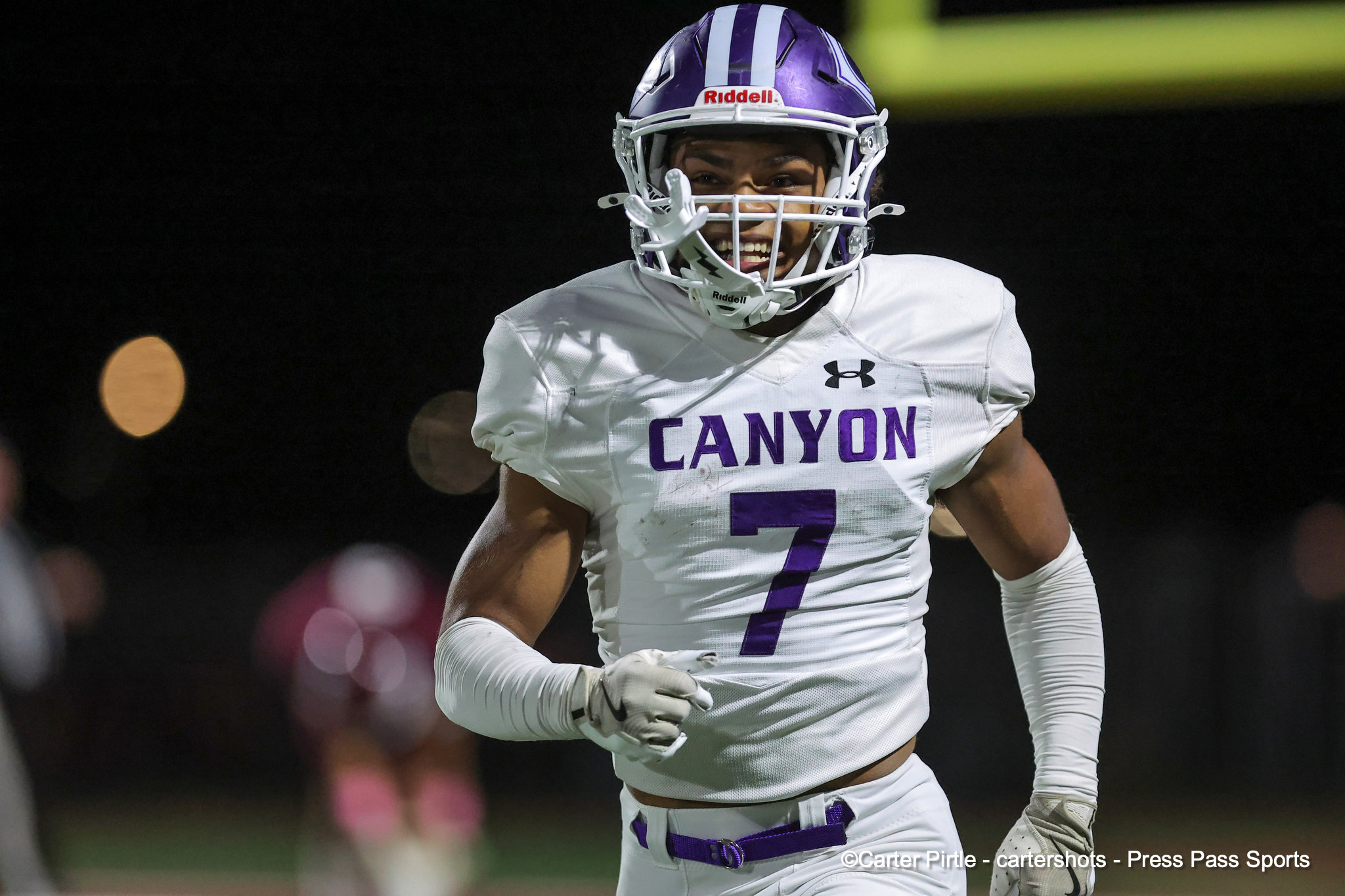 Football player in a white and purple Canyon jersey (#7) running on the field at night, wearing a purple-striped helmet and white facemask, smiling.