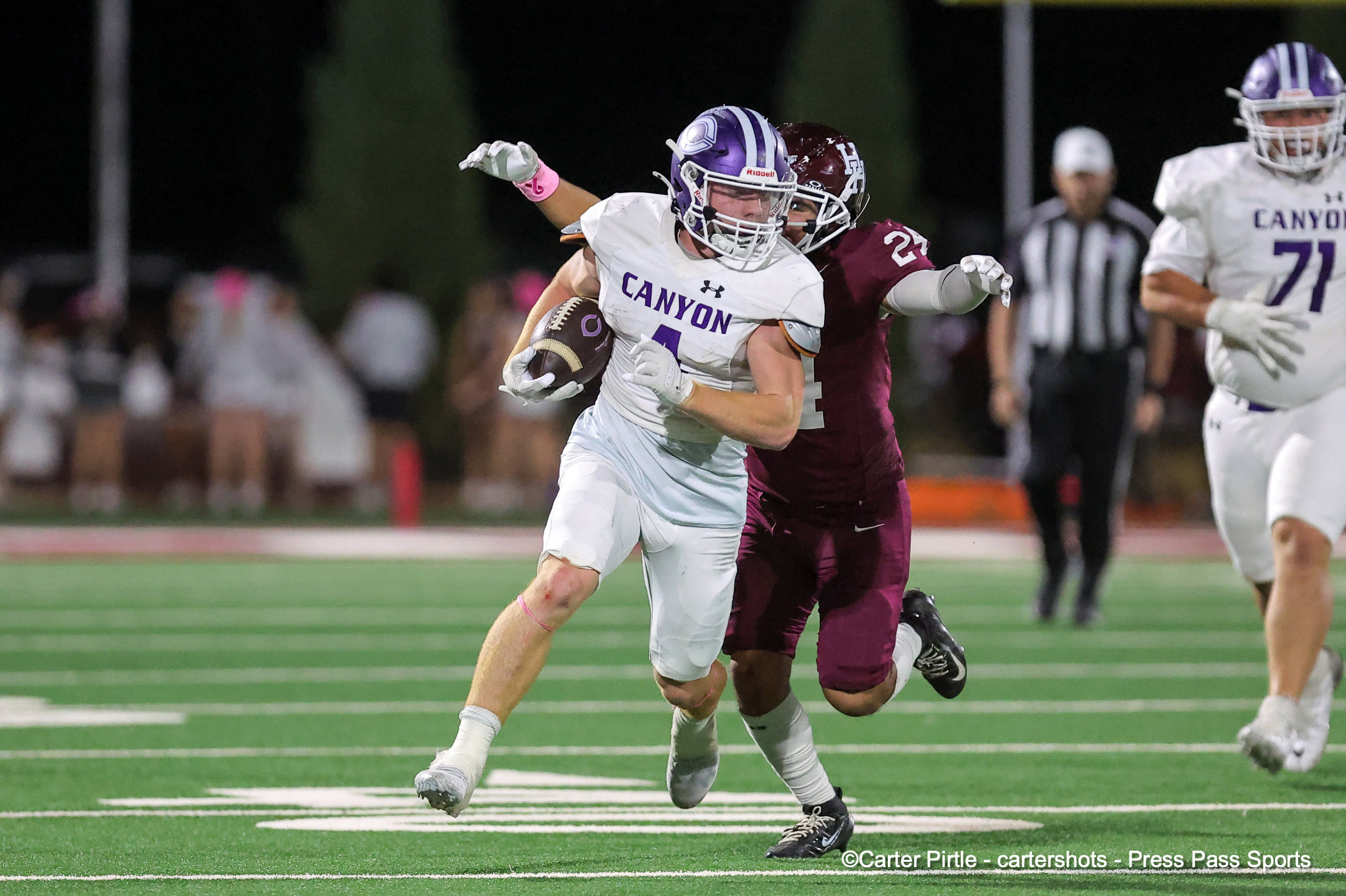 Canyon player in white jersey with the ball, pursued by a maroon-clad defender during a nighttime football game.