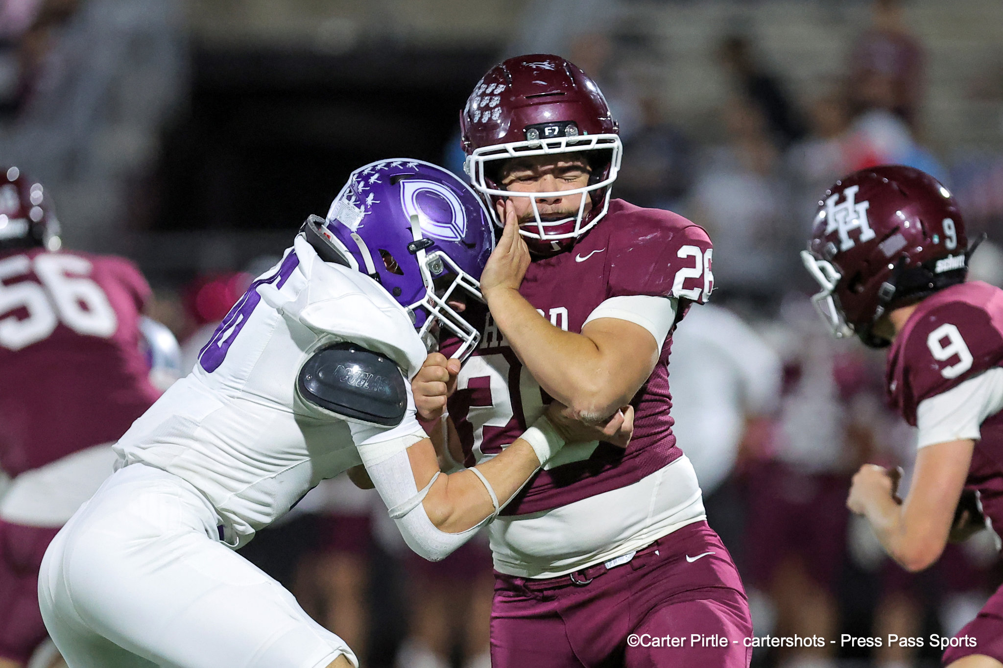 Two football players clash in a tackle during a game; one in maroon jersey number 26 with white helmet faces off against a white-uniform player with a purple helmet.