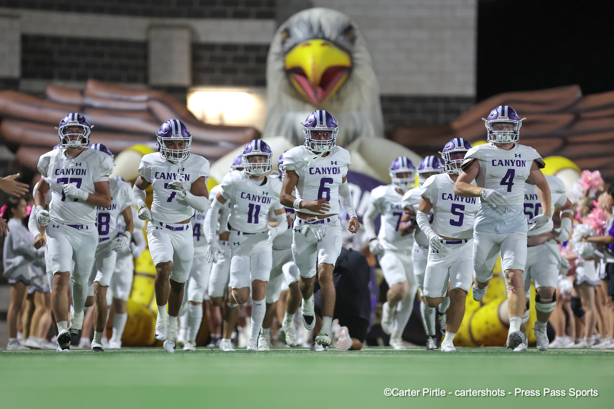 Canyon football players run onto the field in white uniforms with purple numbers as a large eagle mascot looms in the background.