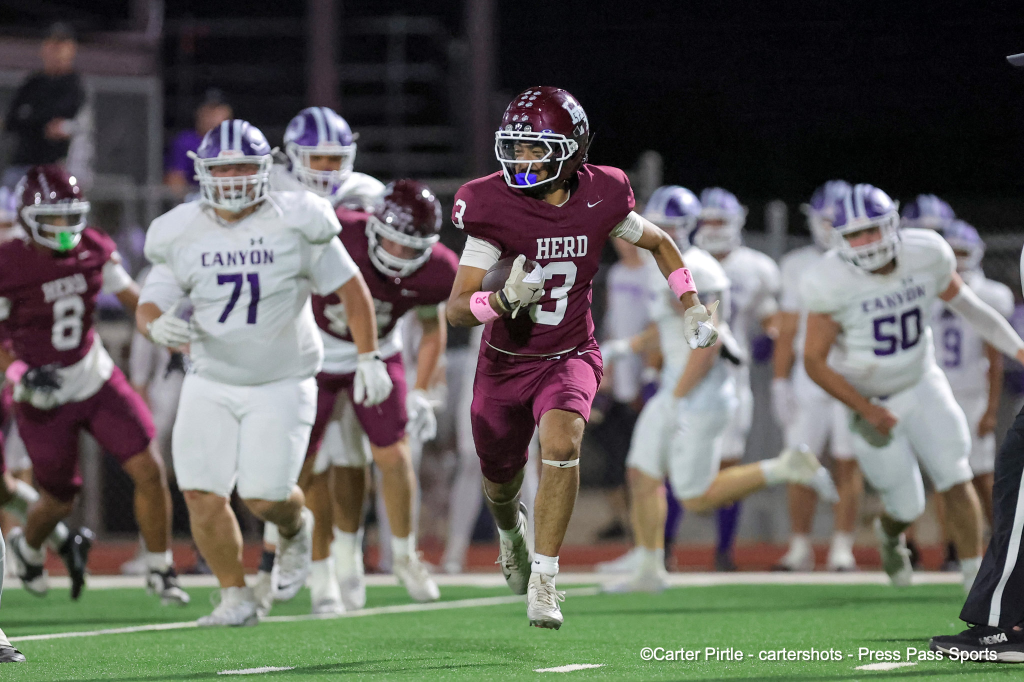 Male football player in maroon HERD jersey runs with the ball during a night game, pursued by opponents in white Canyon uniforms on a green field.