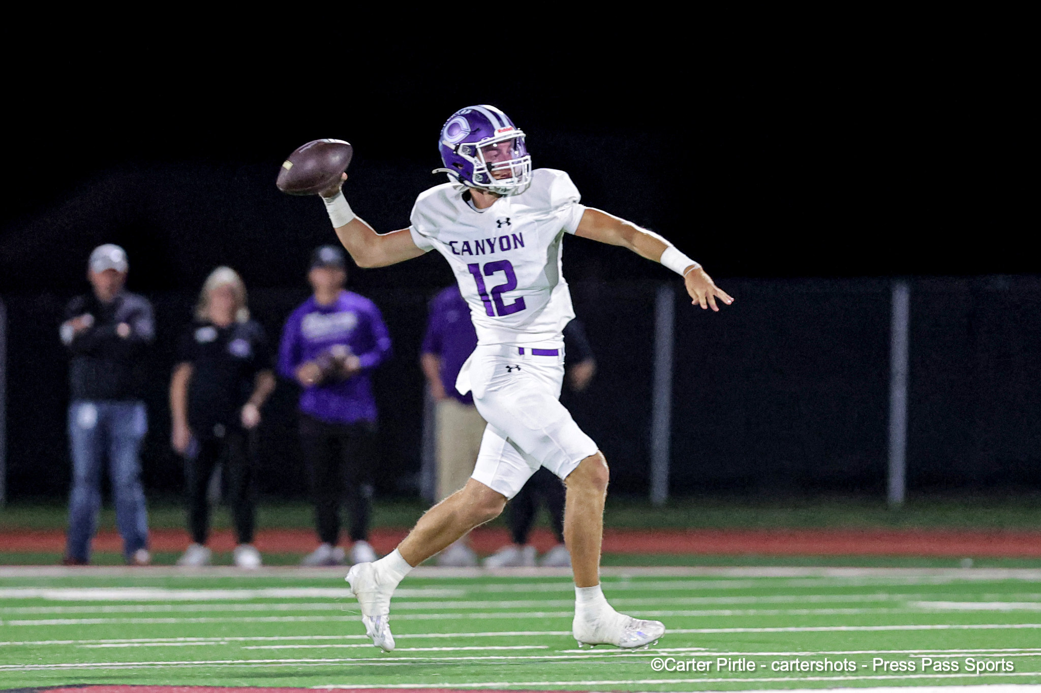 Quarterback in white Canyon jersey #12 throws a pass under stadium lights with spectators in the background on a night game.