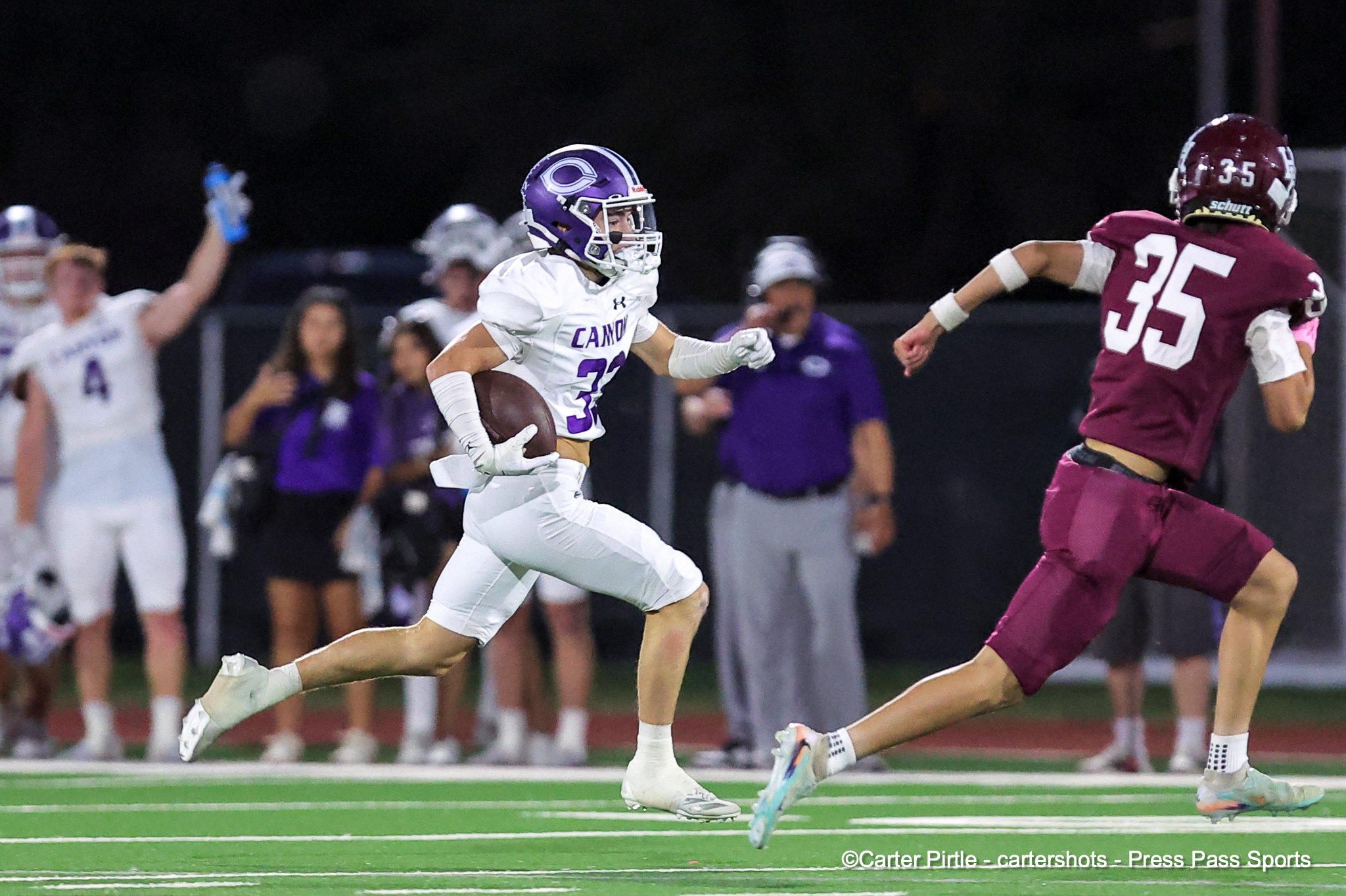 White-uniformed football player carrying the ball, number 32, sprinting downfield as a maroon-clad defender (number 35) pursues him on a night game.