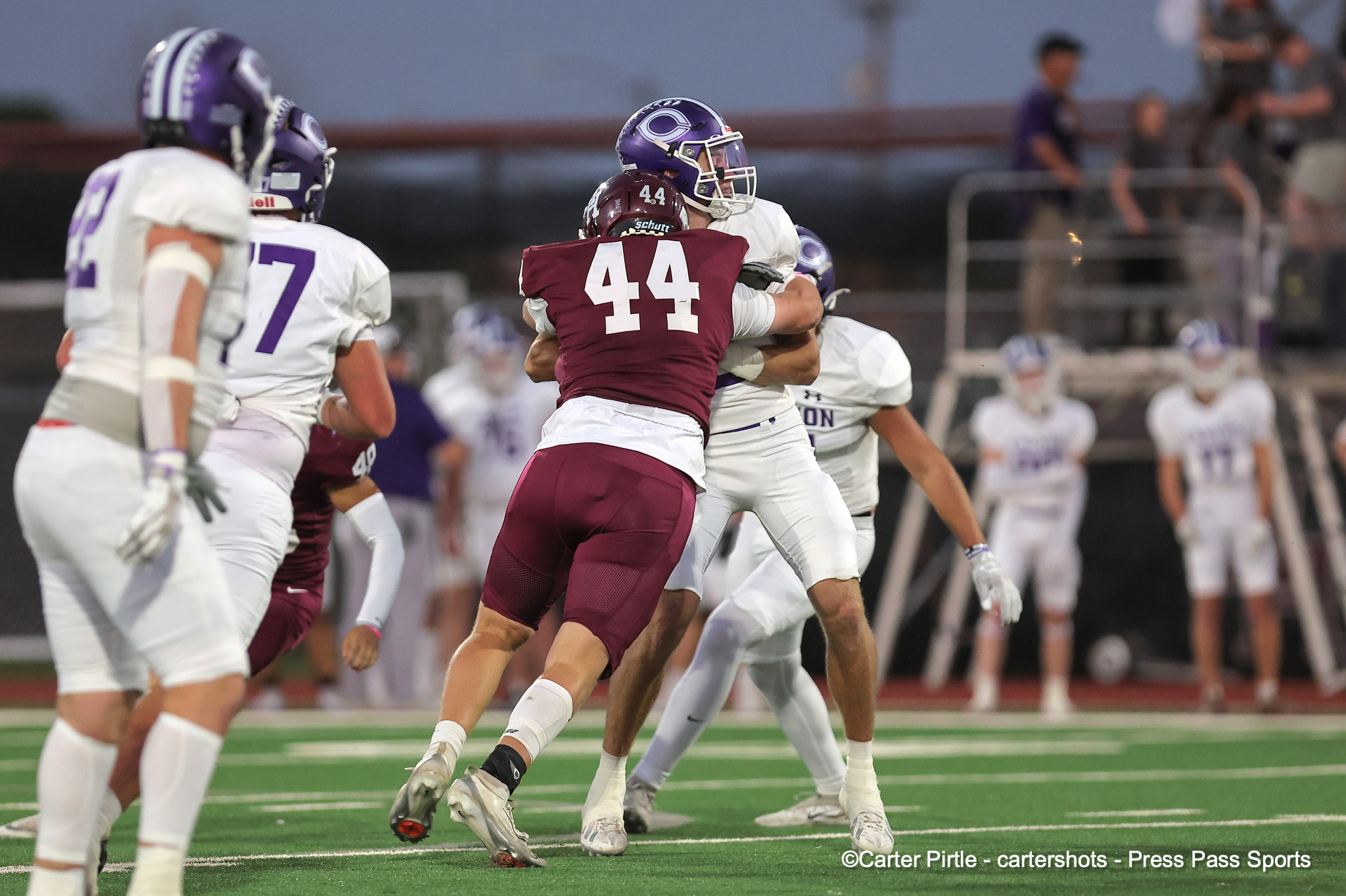 Maroon jersey #44 tackles a white-uniform opponent during a football game on a green turf field.