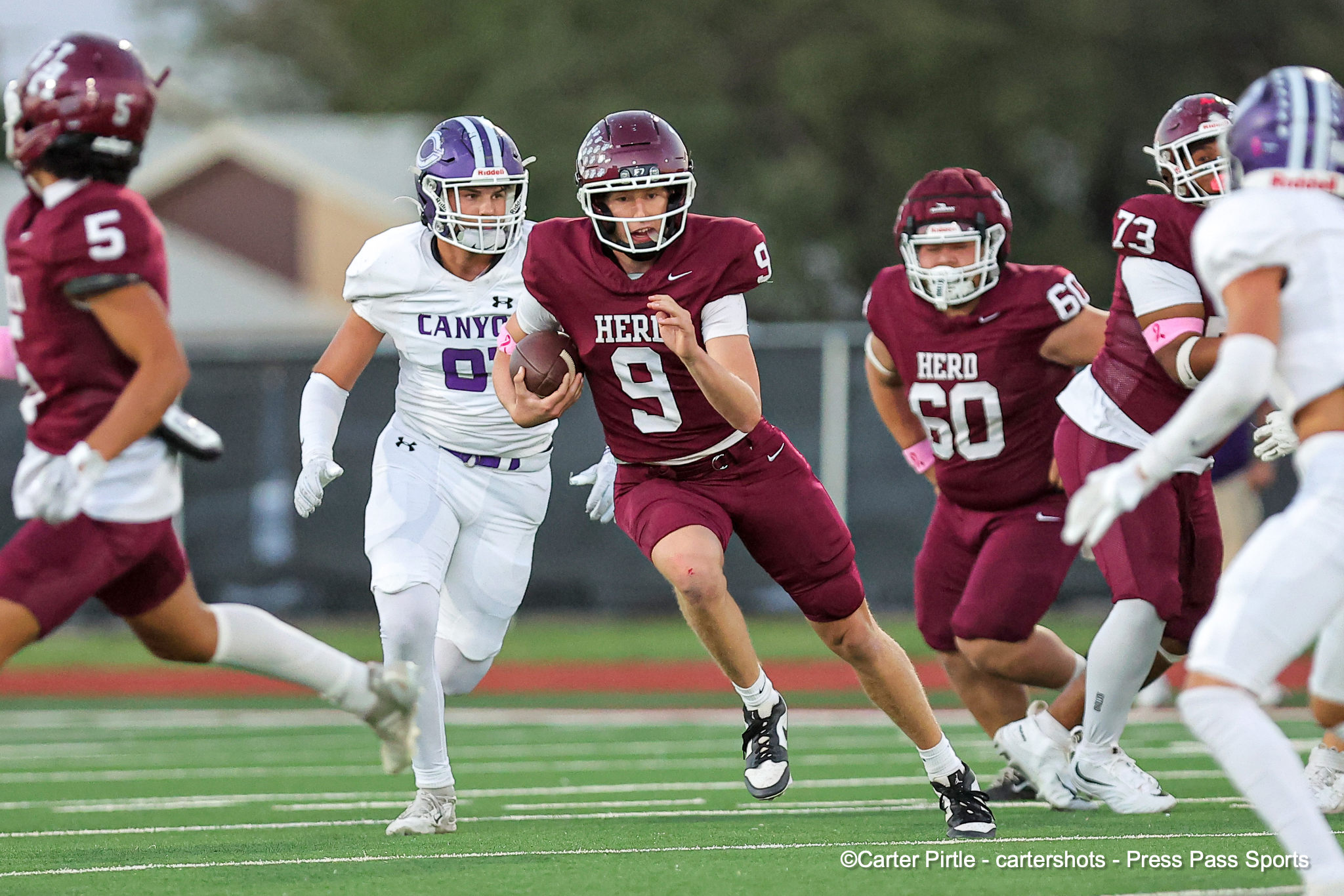 Maroon jerseyed running back (No. 9) carrying the football, pursued by white-jersey defenders on a green football field.