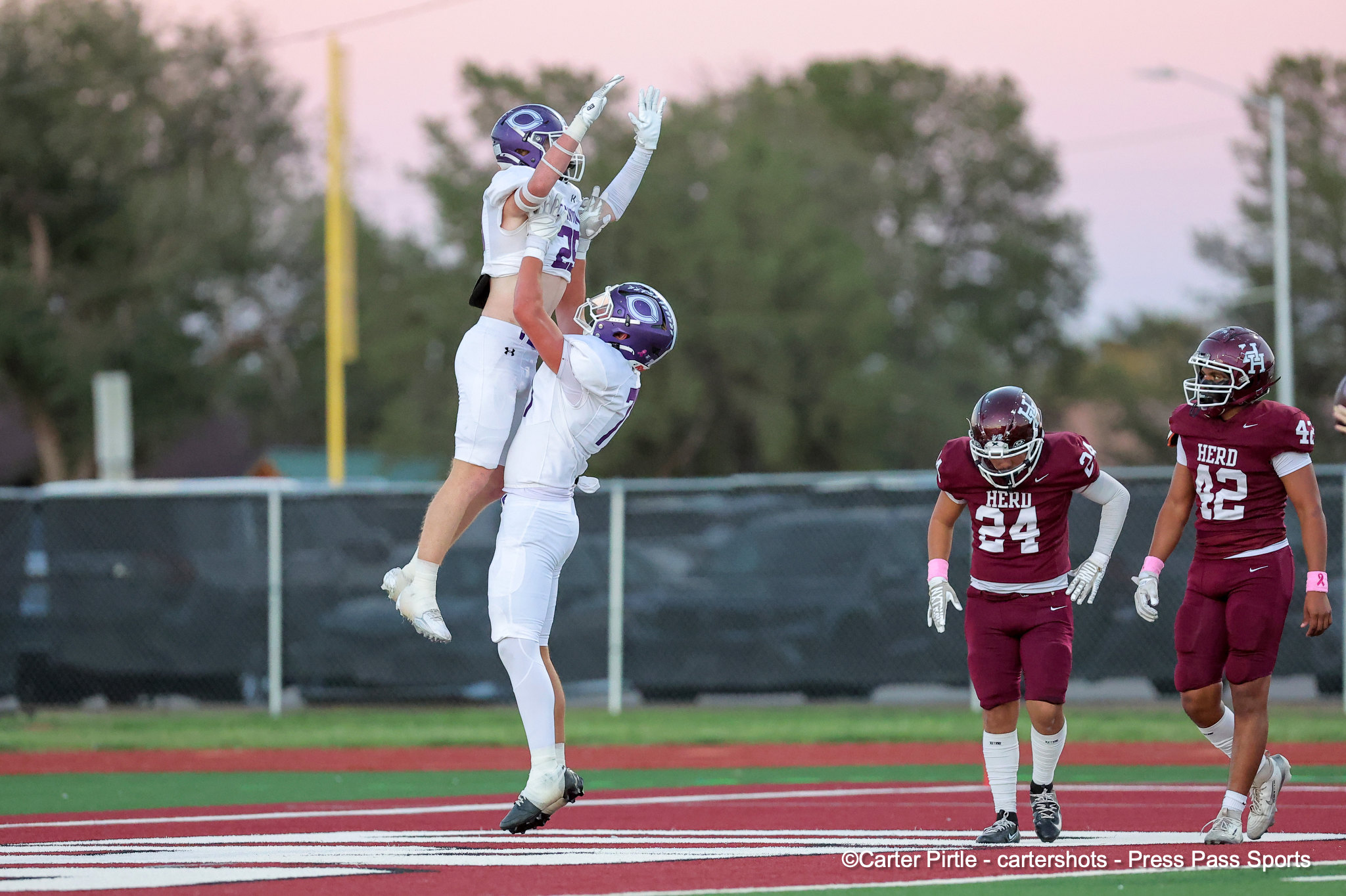 Two football players in white uniforms leap to contest a pass as maroon-clad opponents watch nearby on the field.
