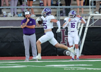 20261009-Canyon-Hereford-FB-015 | Press Pass Sports Football players in white and purple uniforms on the sidelines; player 25 runs with the ball while a teammate (#16) cheers"