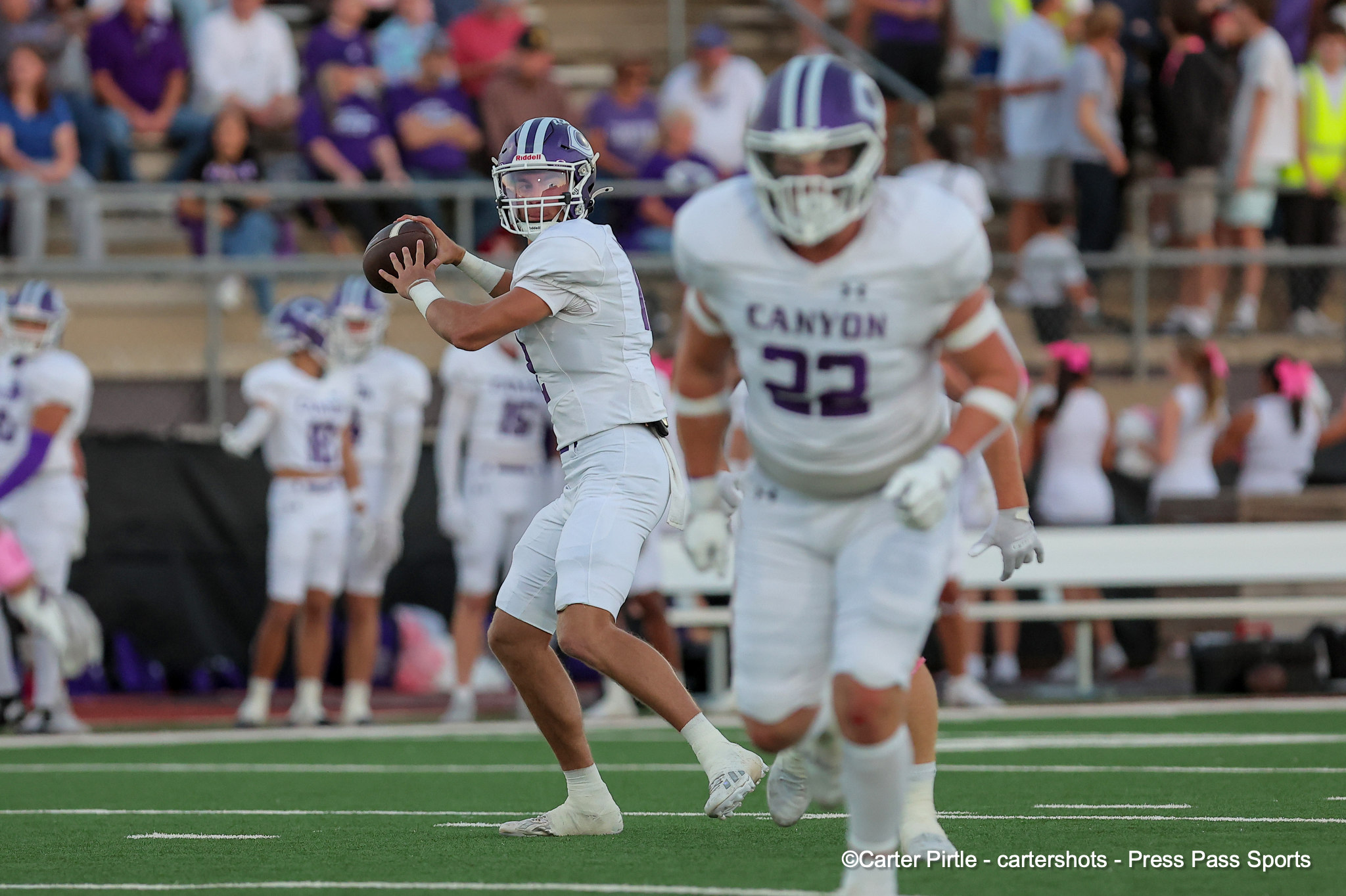 Quarterback in white uniform prepares to pass, holding the ball, with a teammate in front and a cheering crowd in the stands behind on a green football field.