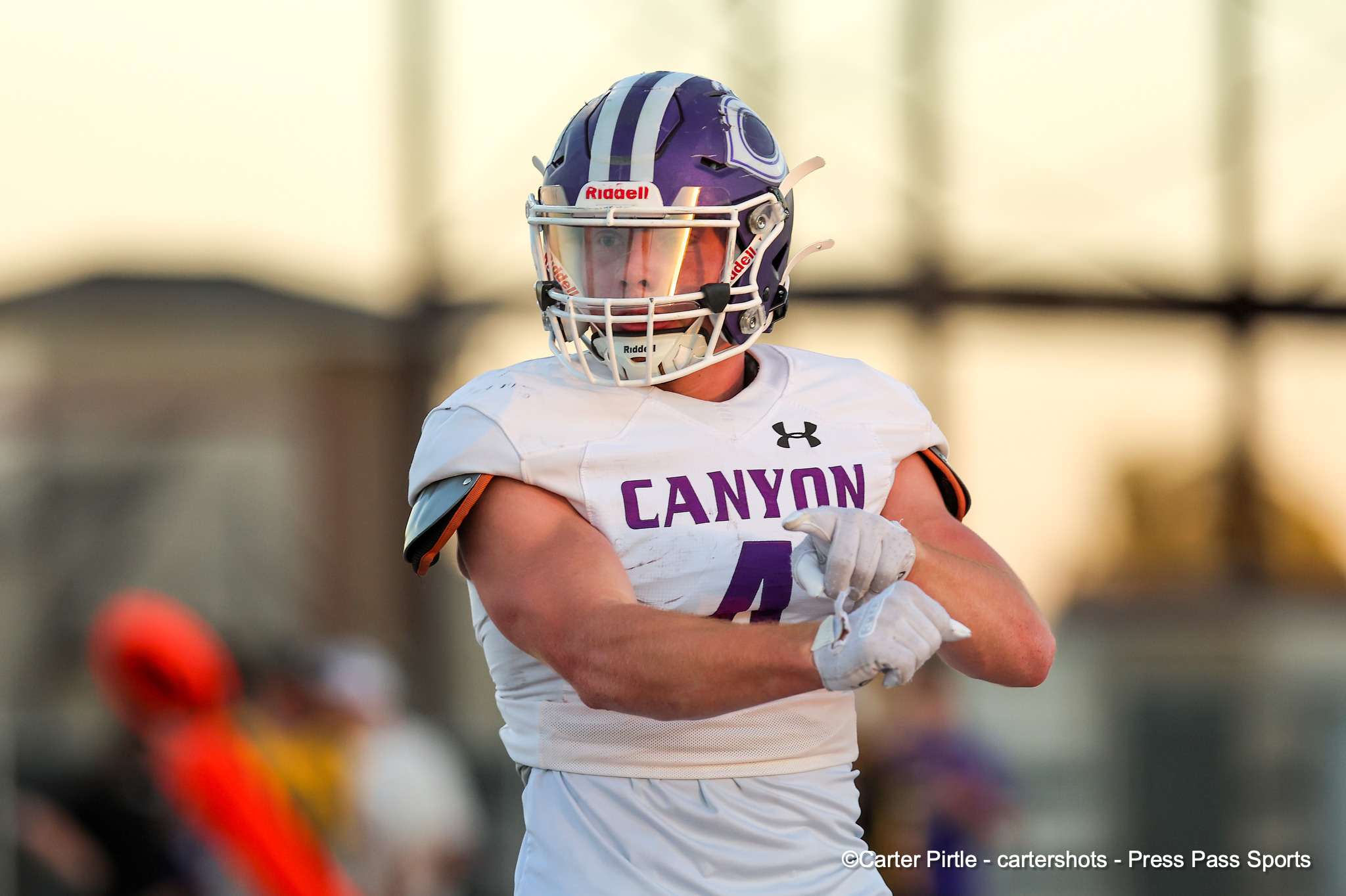 Football player in a white Canyon jersey and purple-striped helmet, mid-action on the field. They wear a faceguard and white gloves.