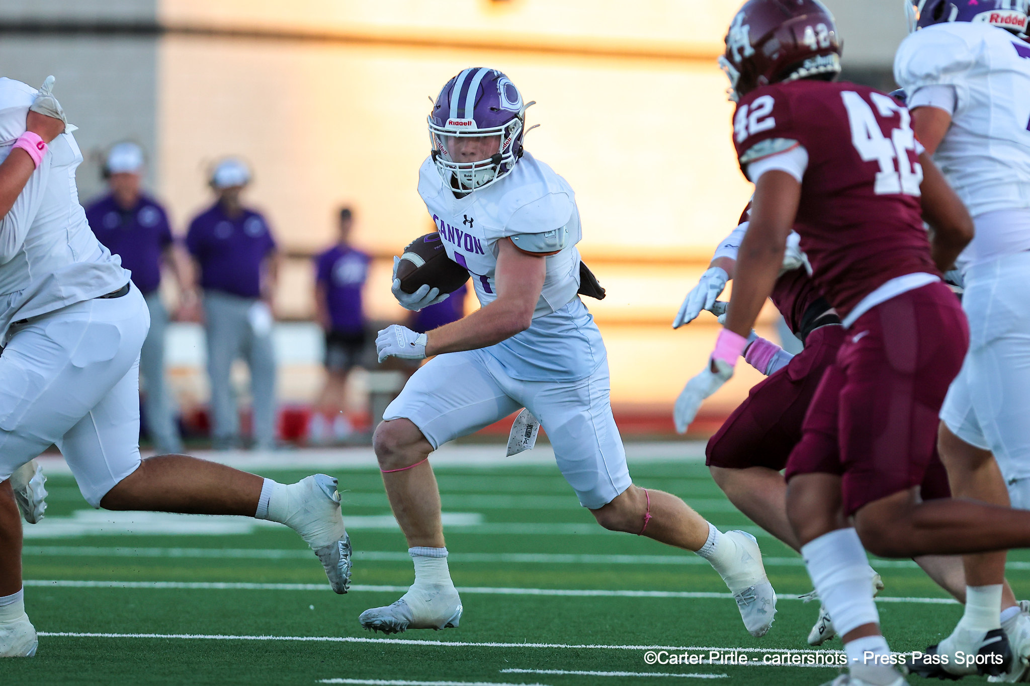 Canyon football player in white jersey and purple helmet runs with the ball as defenders in maroon pursue on a green turf field.