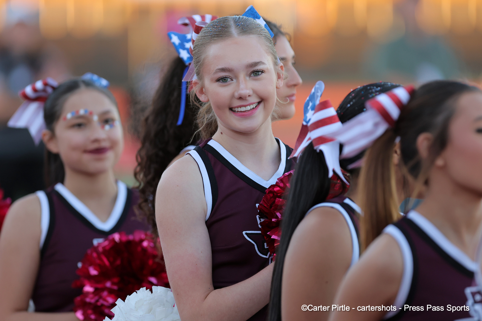 Young cheerleaders in maroon uniforms with red, white, and blue bows, smiling outdoors at sunset, holding pom-poms.