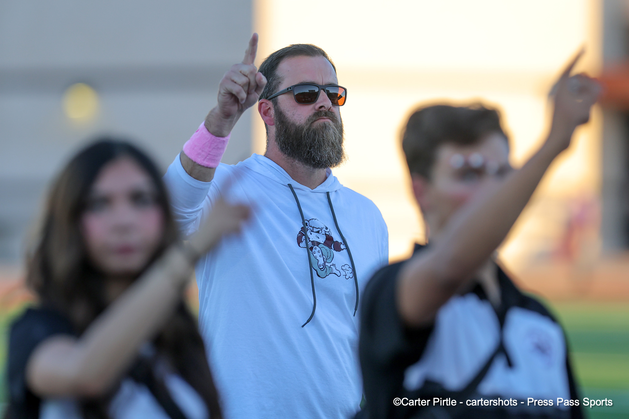 Bearded man in sunglasses and a white hoodie raises his index finger at an outdoor crowd event.