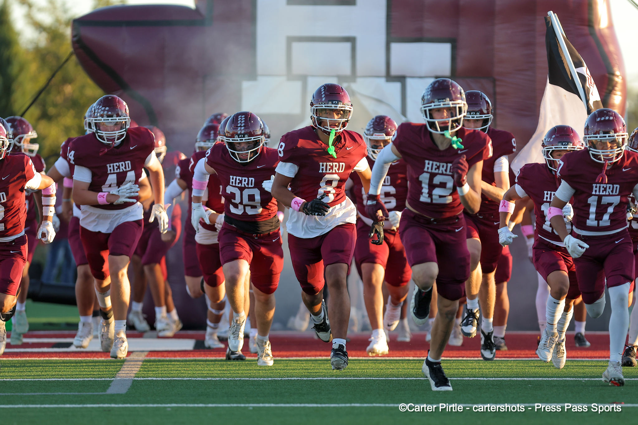 Group of football players in maroon jerseys sprinting onto the field together, helmets on, during a game entrance moment.