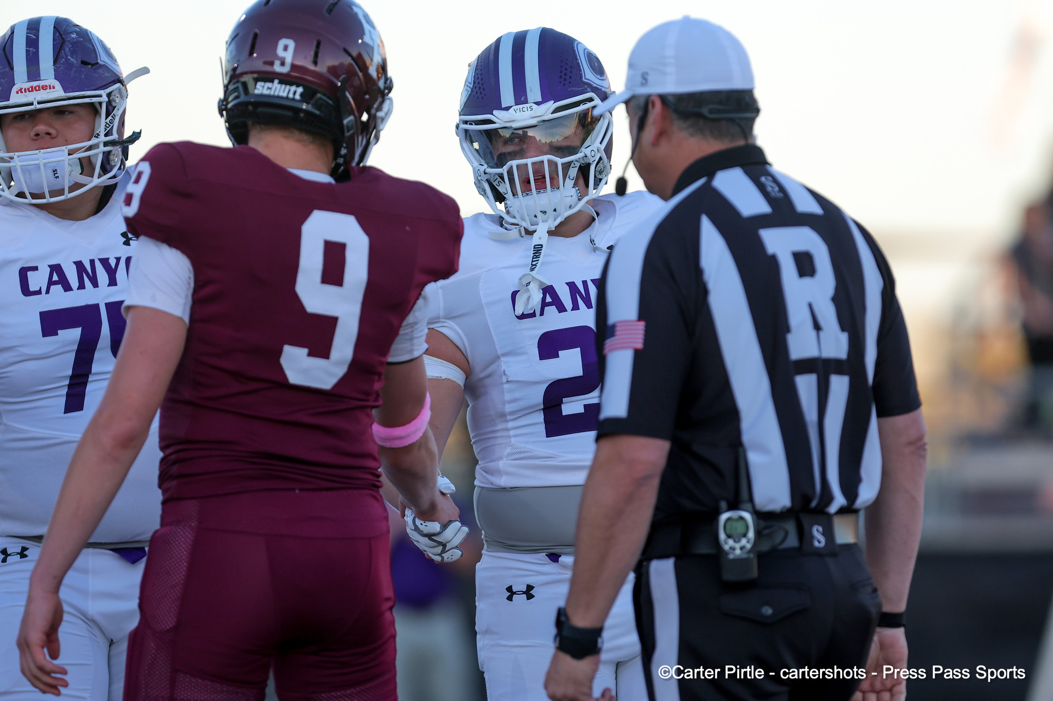 Football players in white and purple uniforms huddle with a maroon-clad opponent and a striped referee nearby.