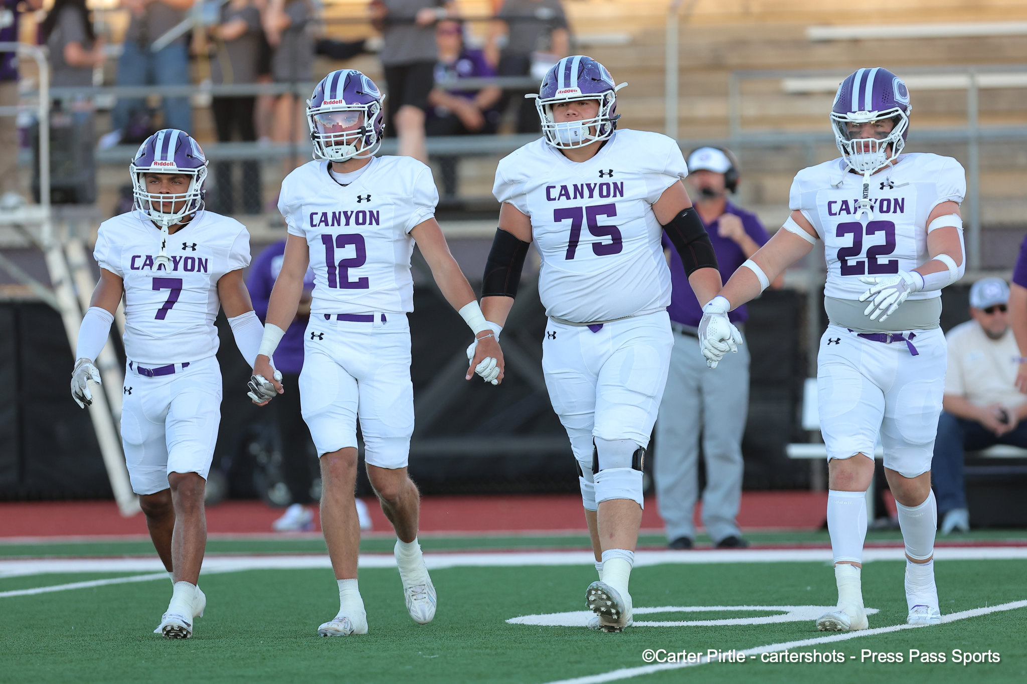 Four Canyon football players in white jerseys and purple helmets walk onto the field together before a game on a green turf."case"