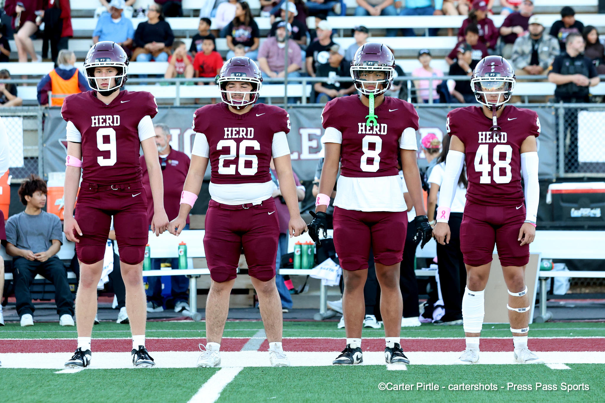 Four football players in maroon jerseys (numbers 9, 26, 8, and 48) stand in a line on the field with a crowd in the bleachers behind them.