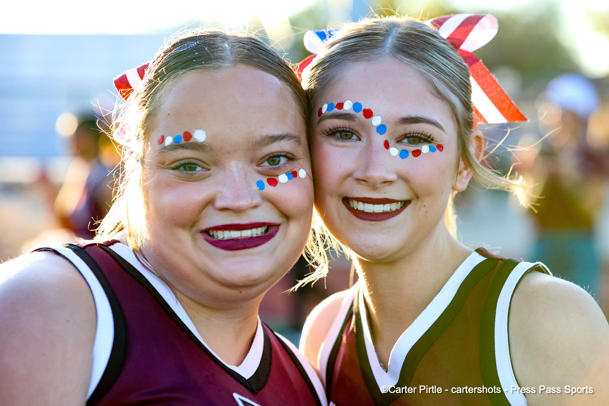 Two smiling cheerleaders posing for a selfie, wearing maroon and olive uniforms with red, white, and blue face paint.