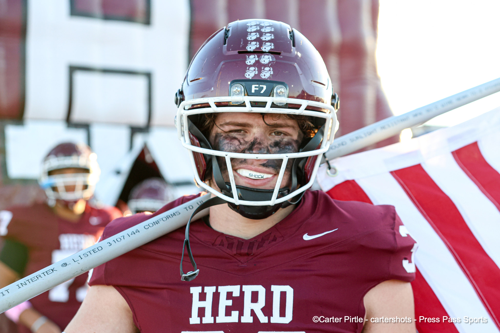 Close-up of a football player in maroon helmet with white face mask, face paint, and a smile, holding a striped banner behind them; jersey reads HERO.