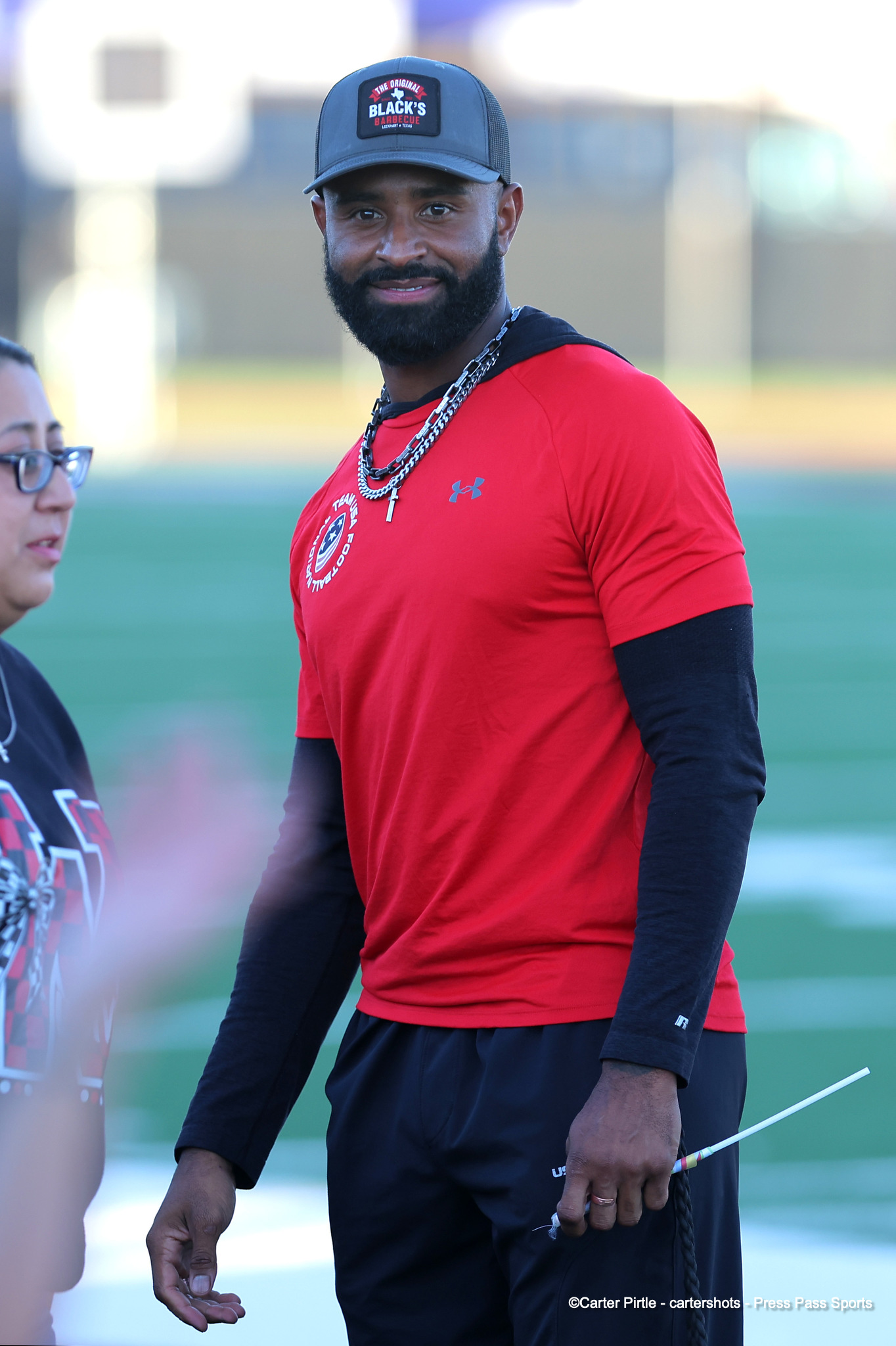 Athlete with a beard in a red athletic shirt and black long sleeves, wearing a cap and a chain, standing on a sports field.
