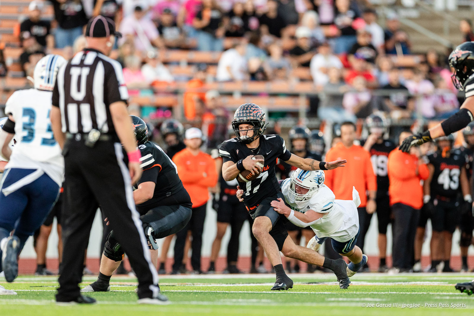 west-plains-vs-dumas-football-jpegJoe-pps-4 | Press Pass Sports Football player in black jersey runs with ball while tackled by a defender in white jersey on a green field; crowd in stands.