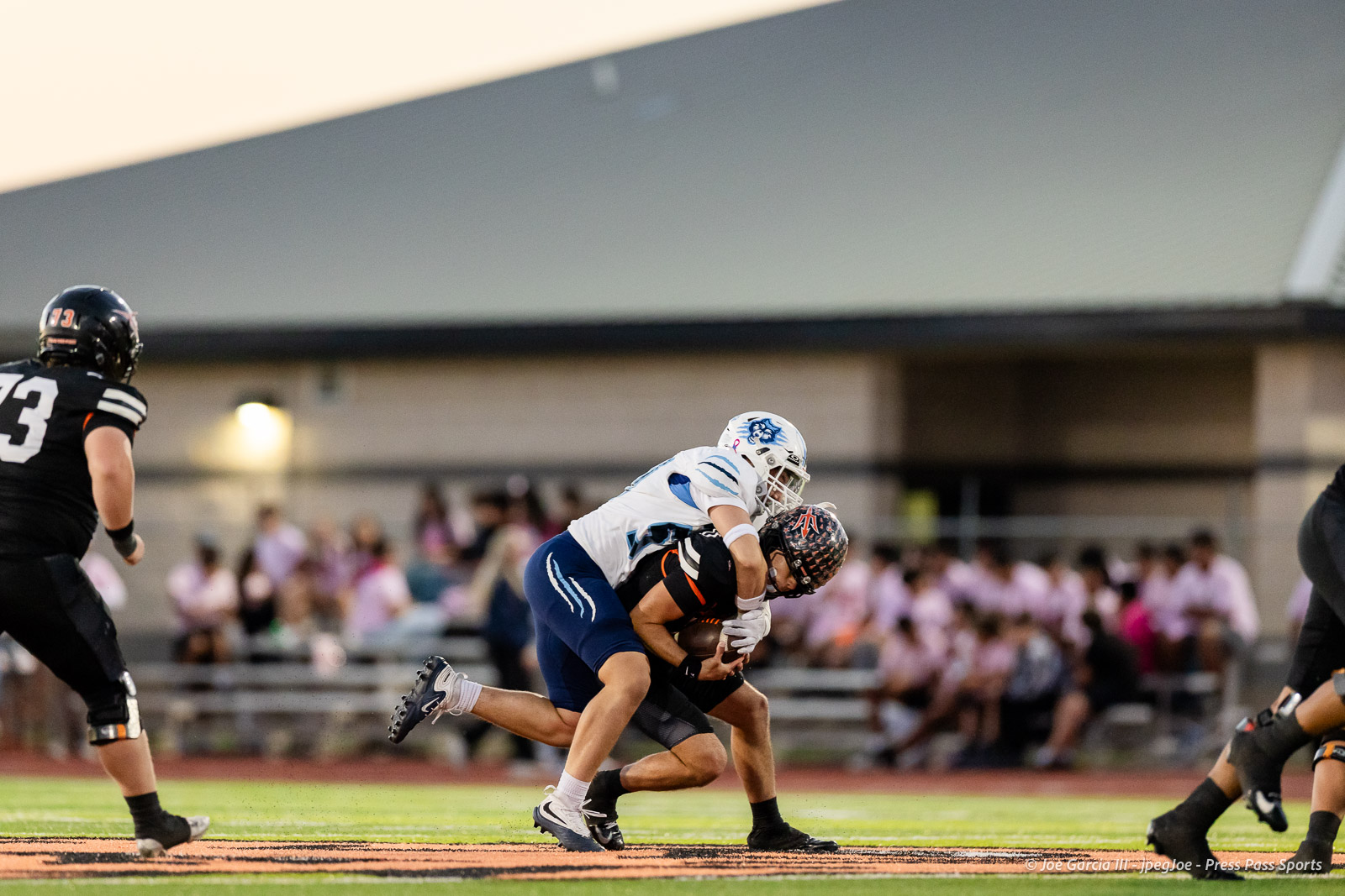 west-plains-vs-dumas-football-jpegJoe-pps-10 | Press Pass Sports Two football players tackle on a green field: a white-and-blue defender grips a black-uniformed ball carrier as crowds watch in the blurred background.