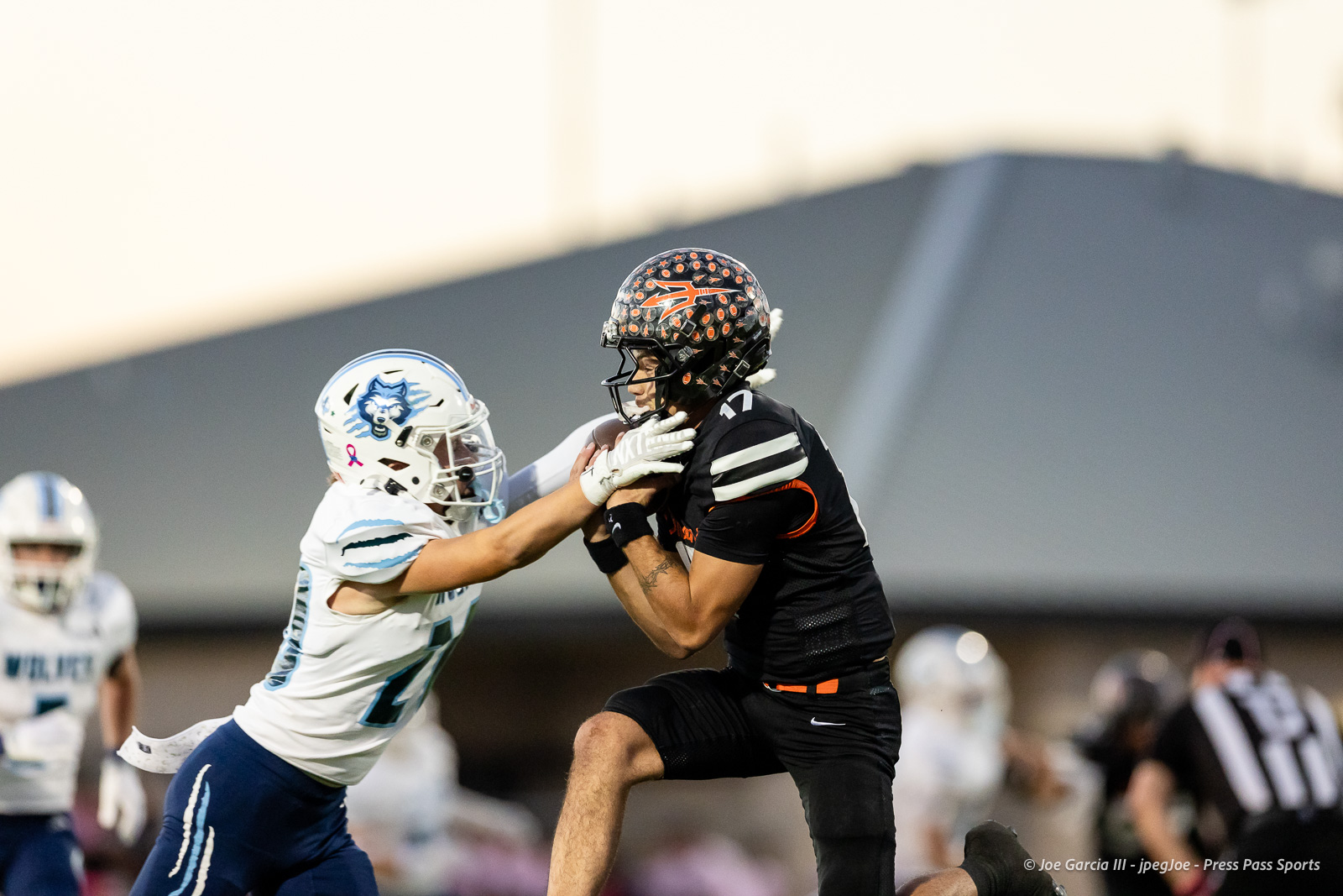 west-plains-vs-dumas-football-jpegJoe-pps-12 | Press Pass Sports Two football players collide mid-air: a black-uniform player with the ball is tackled by a white-uniform defender reaching in front of him, on a blurred stadium backdrop.
