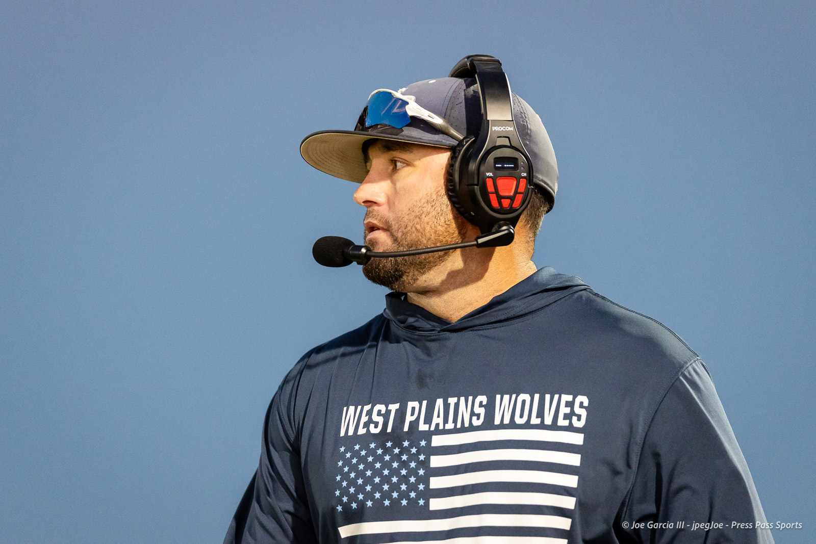 west-plains-vs-dumas-football-jpegJoe-pps-17 | Press Pass Sports Announcer with a headset and microphone, wearing a'West Plains Wolverines' shirt, against a blue sky.
