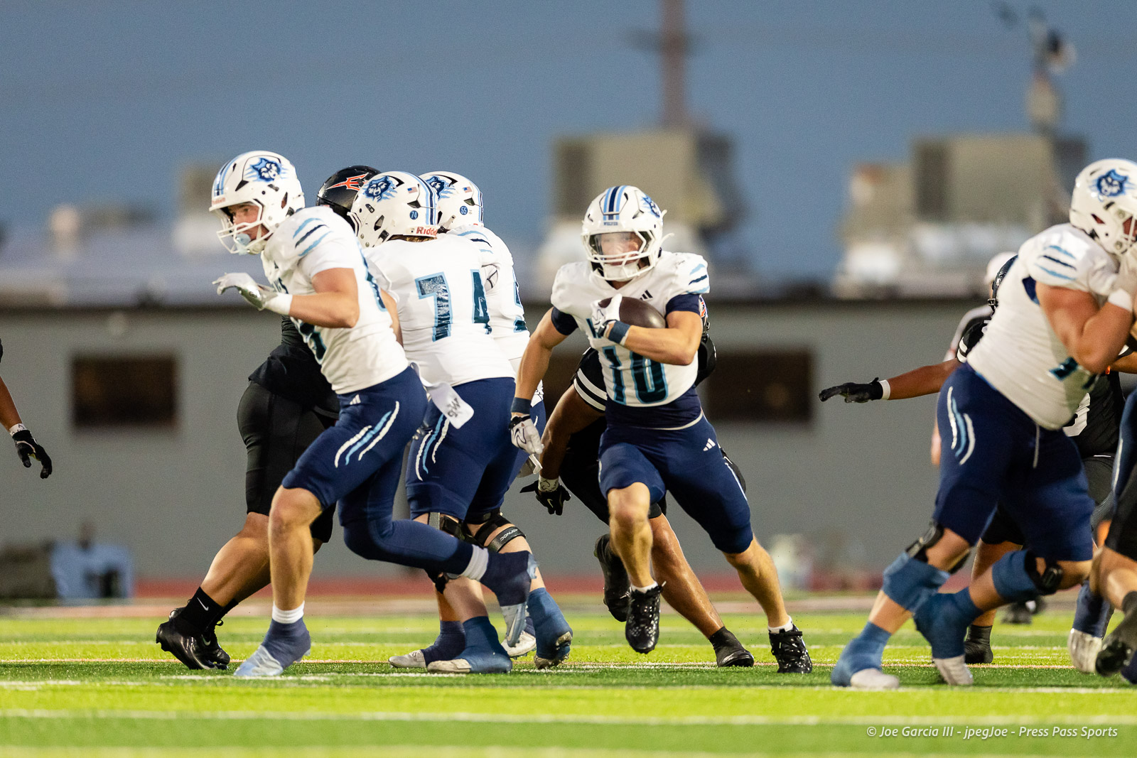 west-plains-vs-dumas-football-jpegJoe-pps-18 | Press Pass Sports Running back with ball in white-and-blue uniform (#10) breaks through a scrum while teammates block and opponents pursue on a bright turf field at dusk
