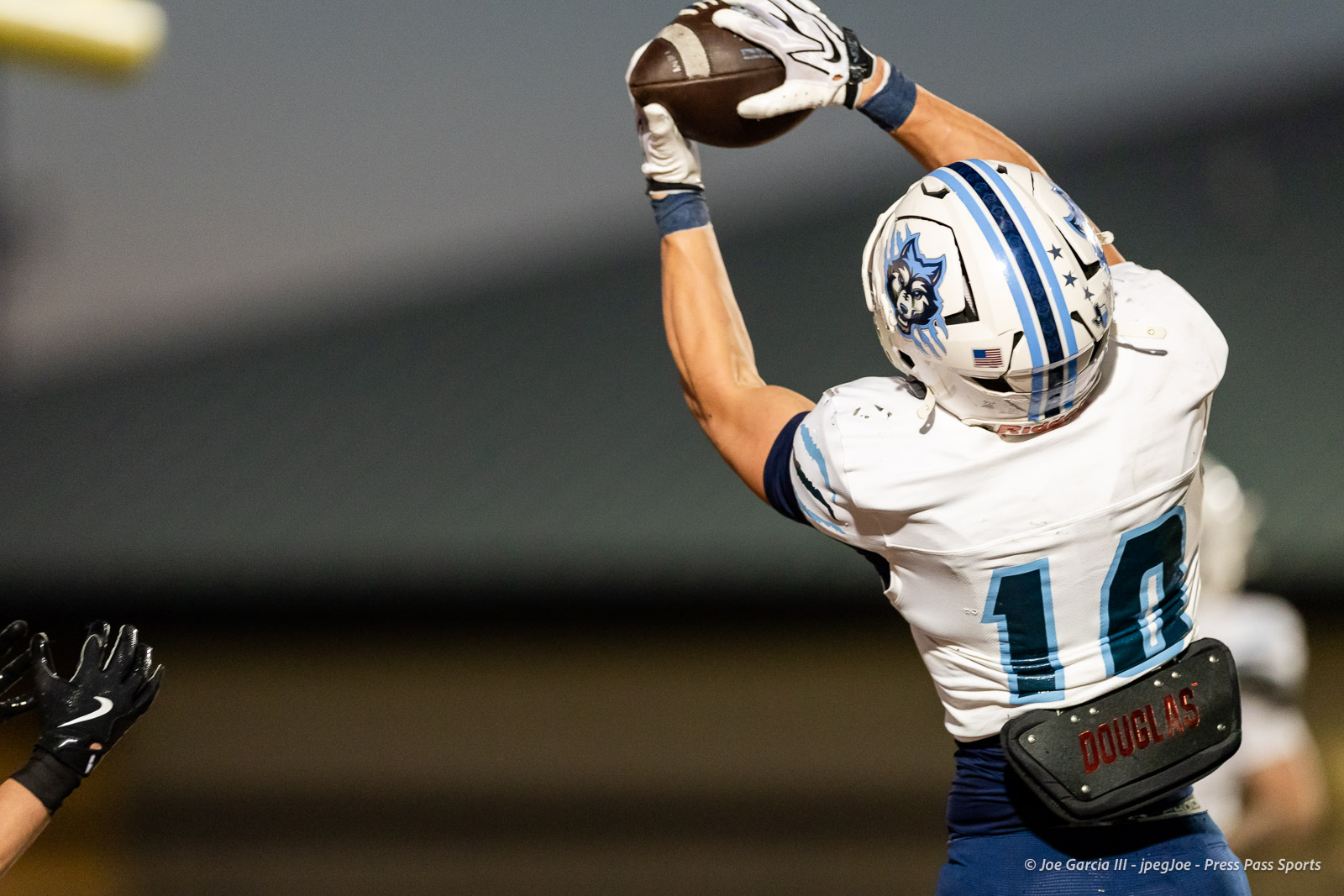 west-plains-vs-dumas-football-jpegJoe-pps-20 | Press Pass Sports Football player in white and blue jersey jumps to catch a football, reaching overhead with gloves, helmet featuring a blue mascot, and a defender's gloved hands in the foreground.